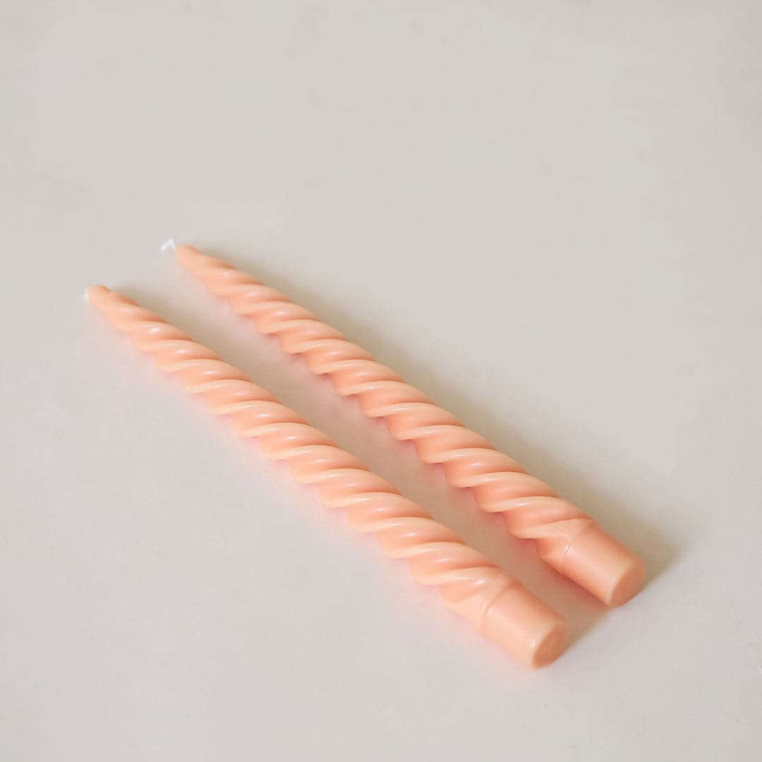 Pair of two pastel pink, twisted taper candles with white cotton wicks lay beside each other against a white background.