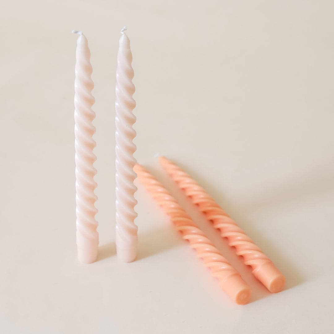 Pair of two blush pink, twisted taper candles stand beside each other. Two pink candles of the same shape lay beside them. 
