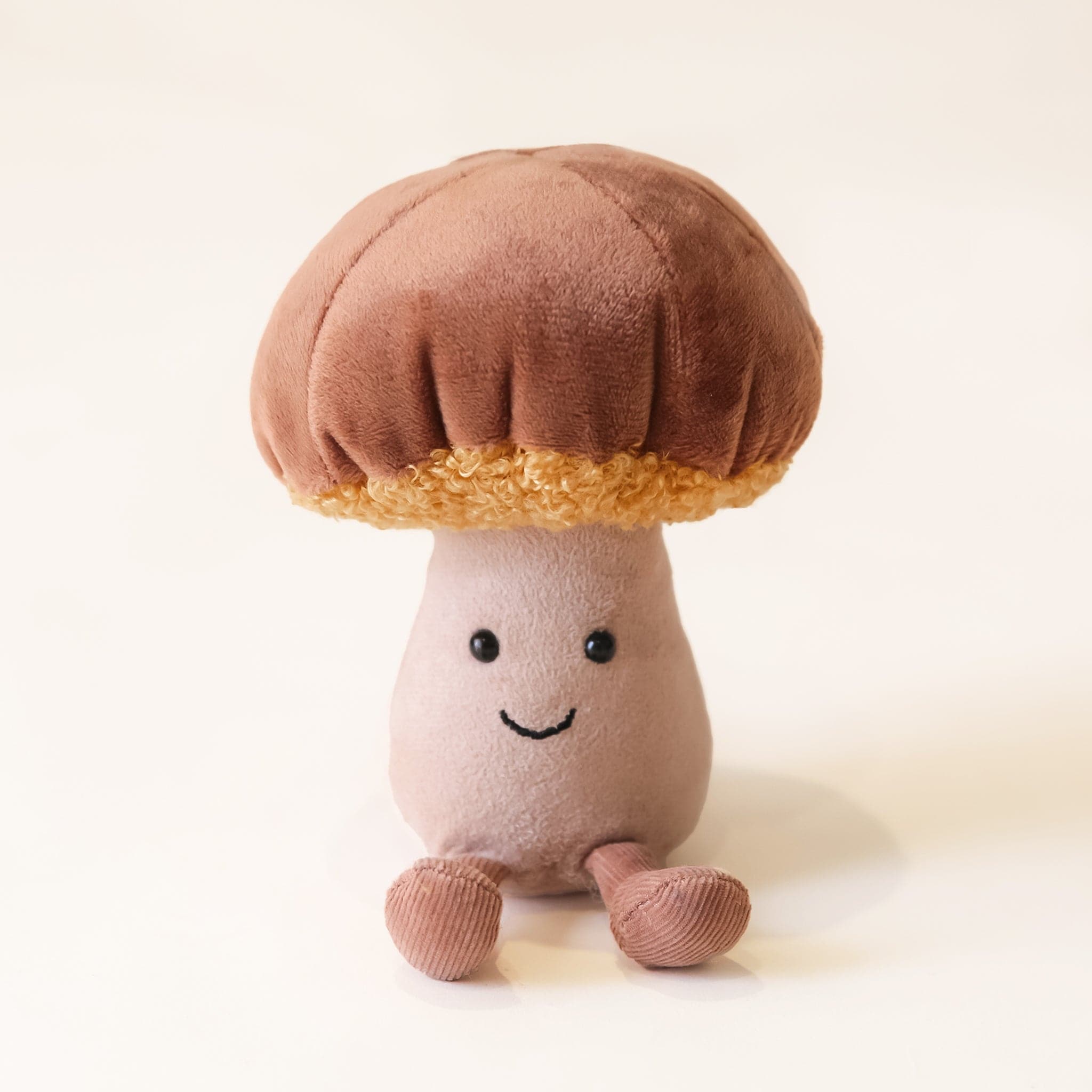 A soft suede stuffed animal in the shape of a mushroom with light brown cap with curly soft gold underneath, a tan colored stalk with smiley face and brown floppy legs.