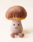 A soft suede stuffed animal in the shape of a mushroom with light brown cap with curly soft gold underneath, a tan colored stalk with smiley face and brown floppy legs.