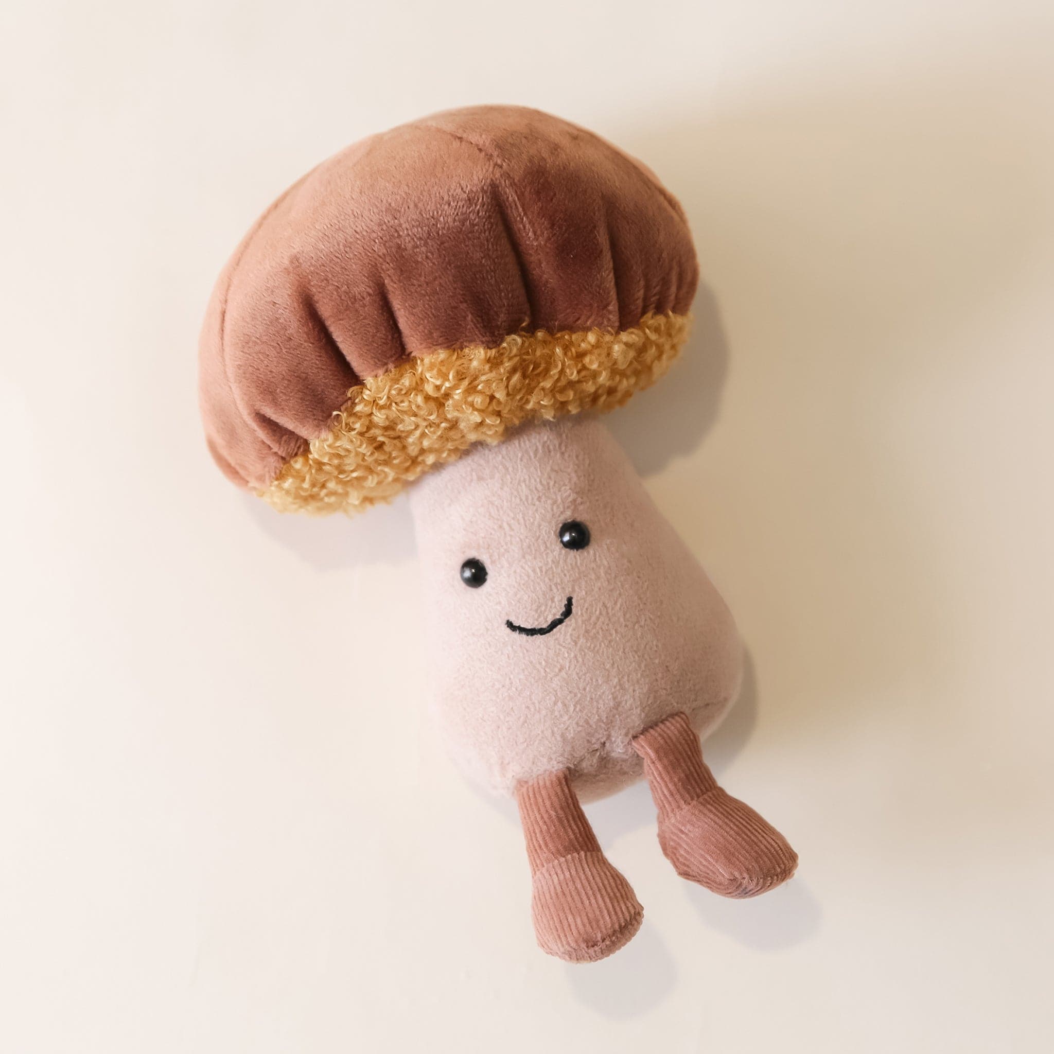 A soft suede stuffed animal in the shape of a mushroom with light brown cap with curly soft gold underneath, a tan colored stalk with smiley face and brown floppy legs.