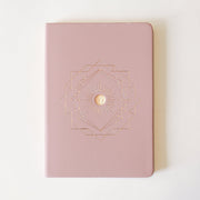 On a cream background is a light pink journal with a geometric gold design in the center. 