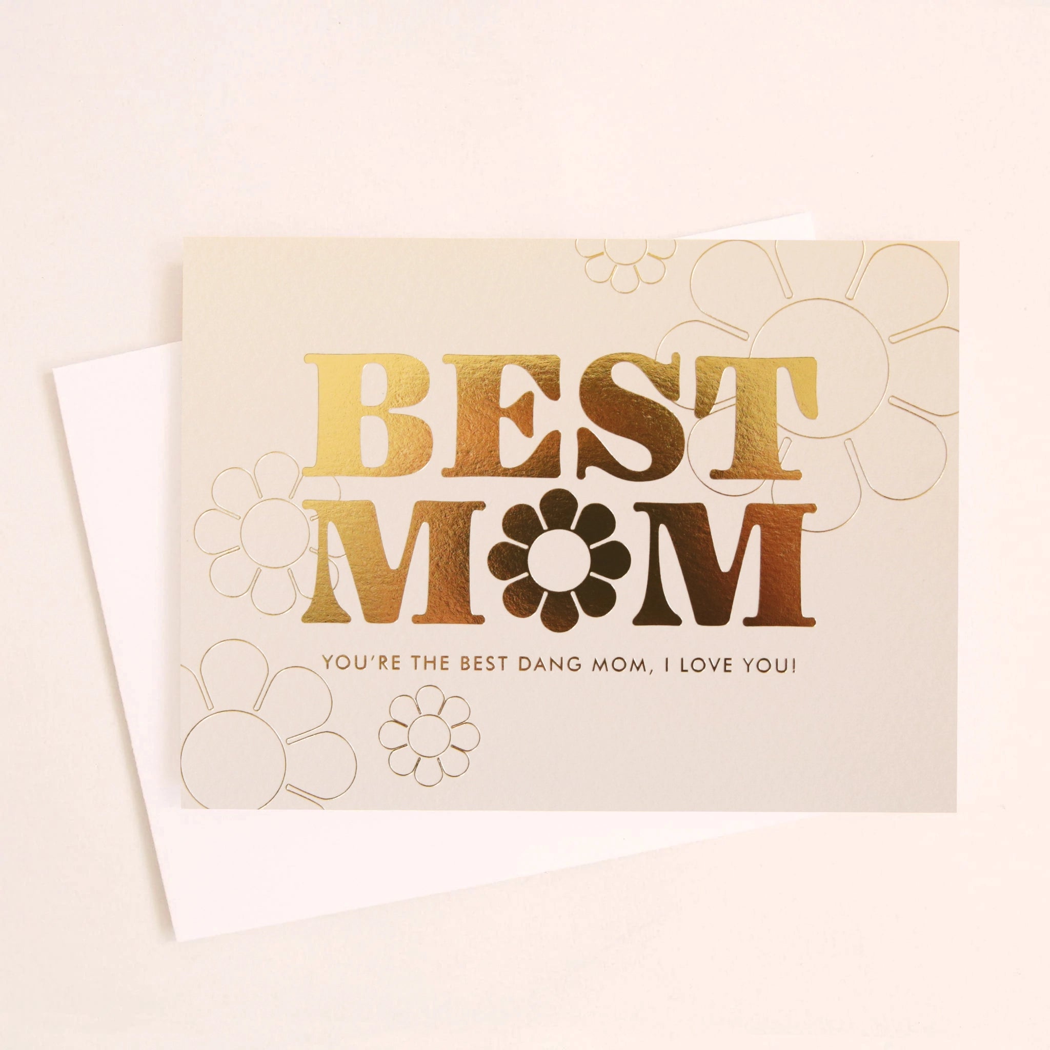 A cream colored card with gold foil lettering that reads, "Best Mom, You're The Best Dang Mom, I Love You!". The 'o' in 'Mom' is a daisy. There are thin gold outlines of daisies in the top right corner and the bottom left. The card is photographed with a coordinating white envelope that is included with purchase.