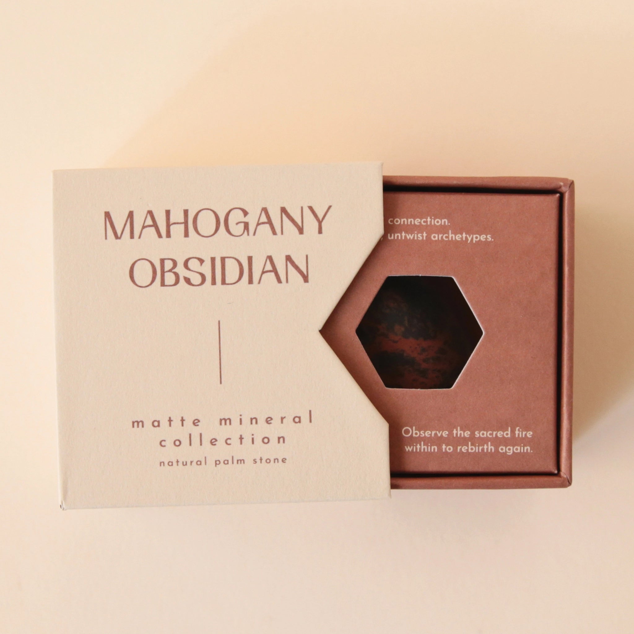 A tan box that reads, "Mahogany Obsidian Matte Mineral Collection" in dark red letters along with a round obsidian stone in front of it that features black and dark red coloring. Keep in mind, each stone is unique and may vary slightly in color and shape.