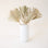 Narrow white cylinder vase filled with neutral toned palm fronds. The vase lays against a blush pink background. 