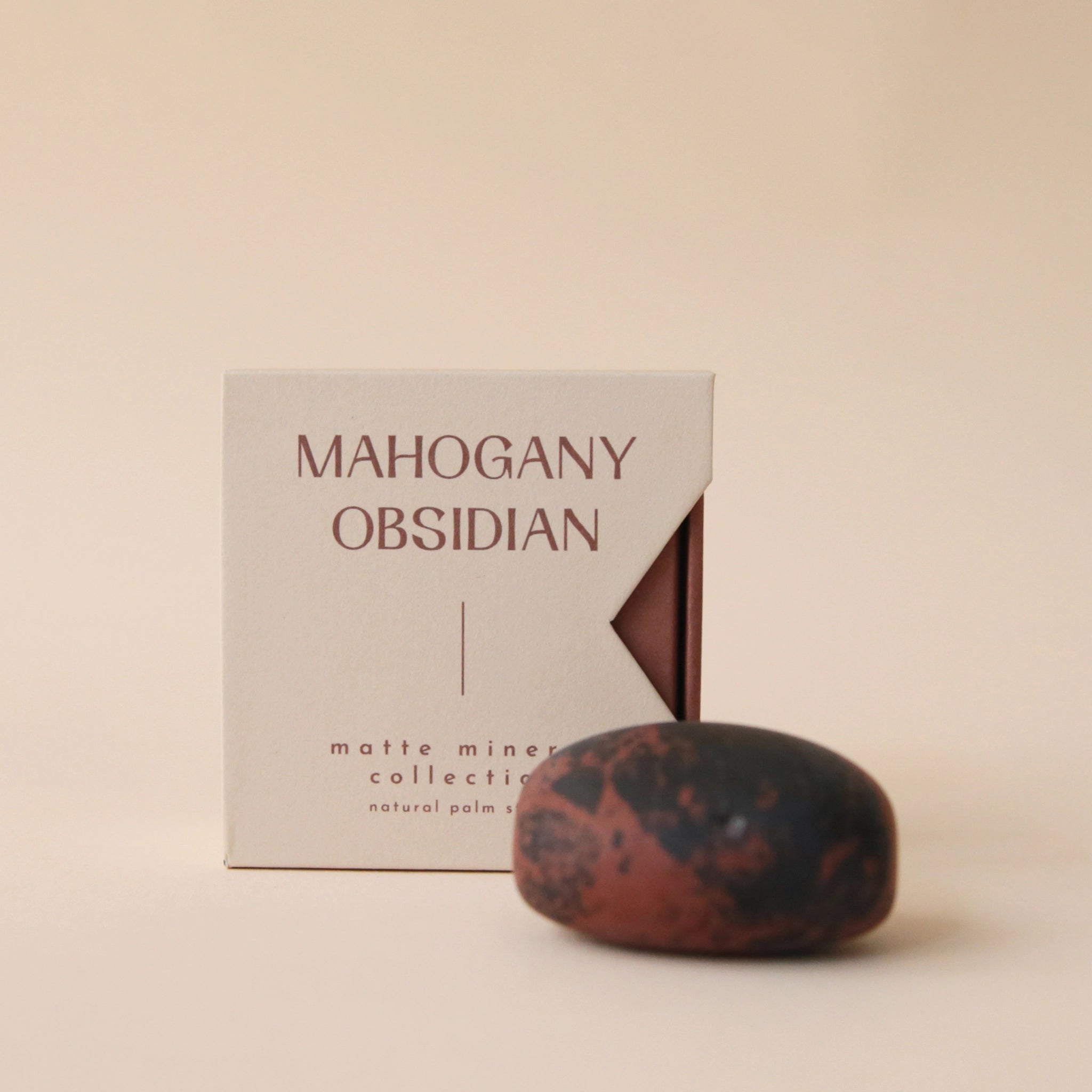 A tan box that reads, &quot;Mahogany Obsidian Matte Mineral Collection&quot; in dark red letters along with a round obsidian stone in front of it that features black and dark red coloring. Keep in mind, each stone is unique and may vary slightly in color and shape.