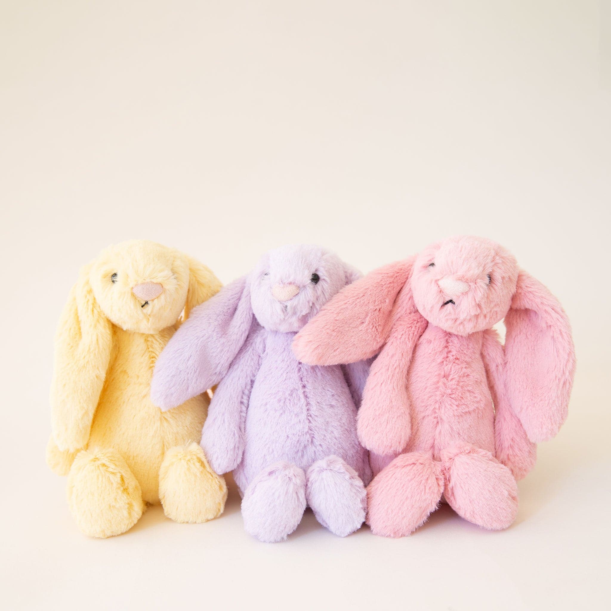 A fuzzy pink bunny stuffed animal with long floppy ears, a light pink nose and black eyes photographed here next to a yellow and lilac version of the bunny also available on our website.