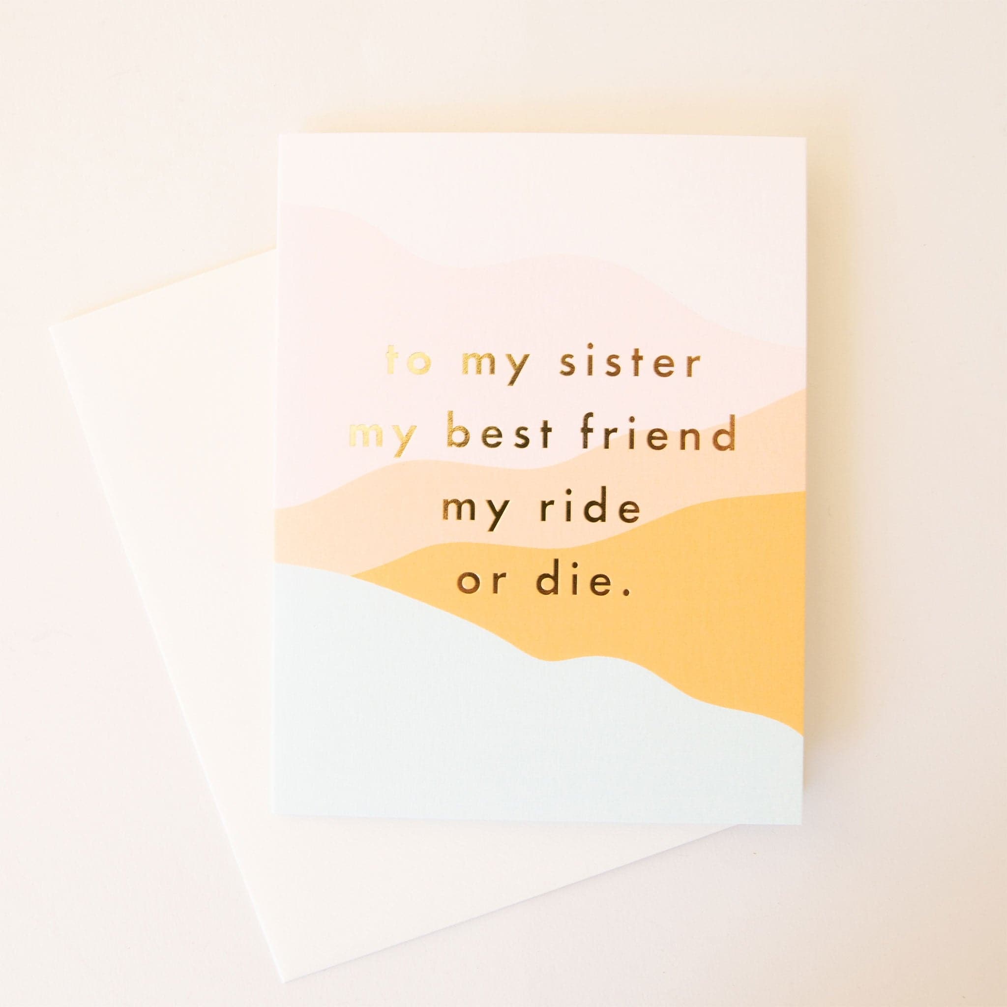 Greeting card reading &#39;to my sister my best friend my ride or die&#39; in gold foil lettering against a soft mosaic mountain background. 