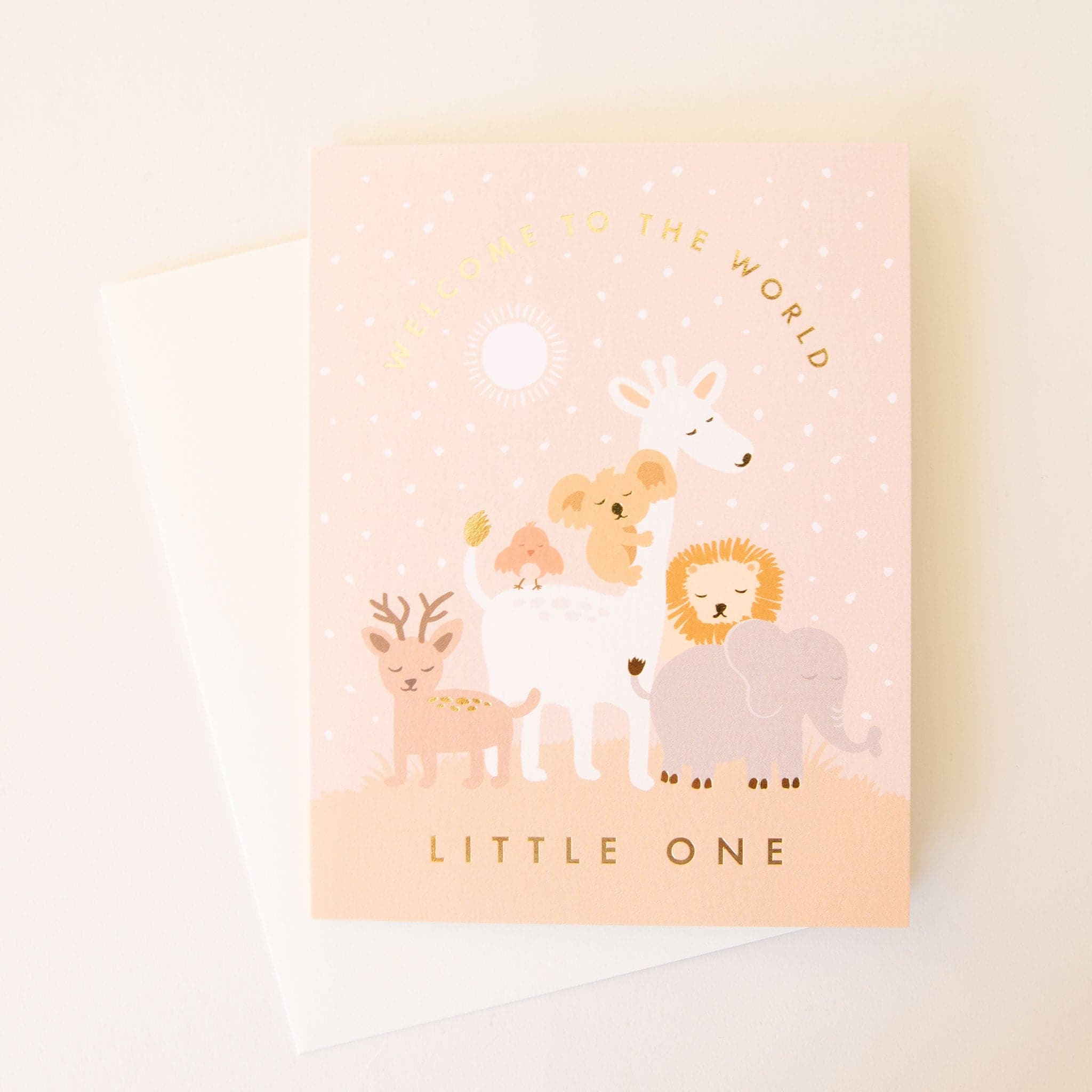 Soft pink card filled with a herd of peaceful animals including a dear, giraffe, koala and more. The text 'Welcome to the world' curves above them in gold foil across a star filled sky. Below the scene reads 'little one' in gold foil capital lettering. The card is accompanied by a solid white envelope. 