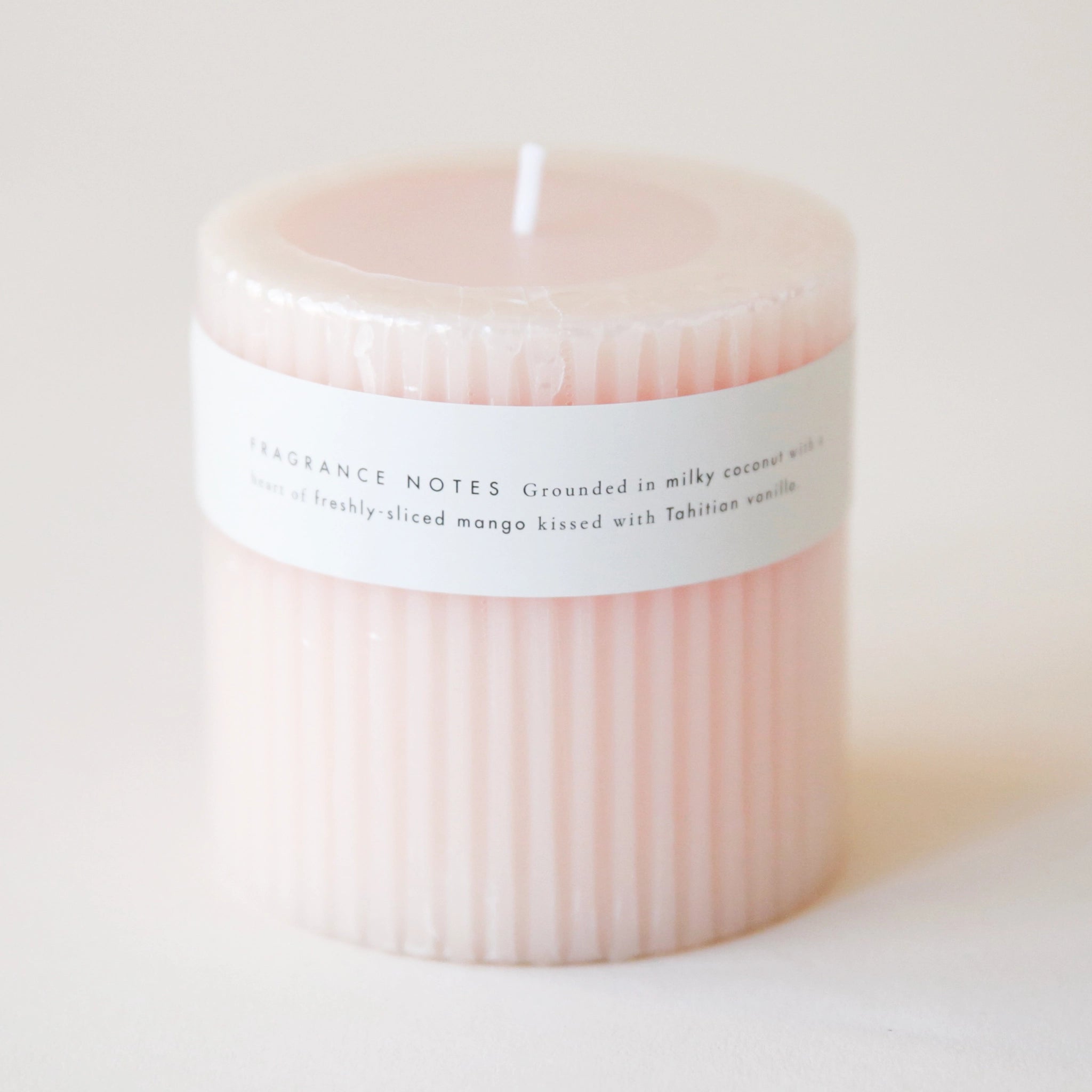 A light pink pilar candle with a ribbed detail and a thin white label wrapped around that reads, "Coconut Milk Mango Scented Candle".