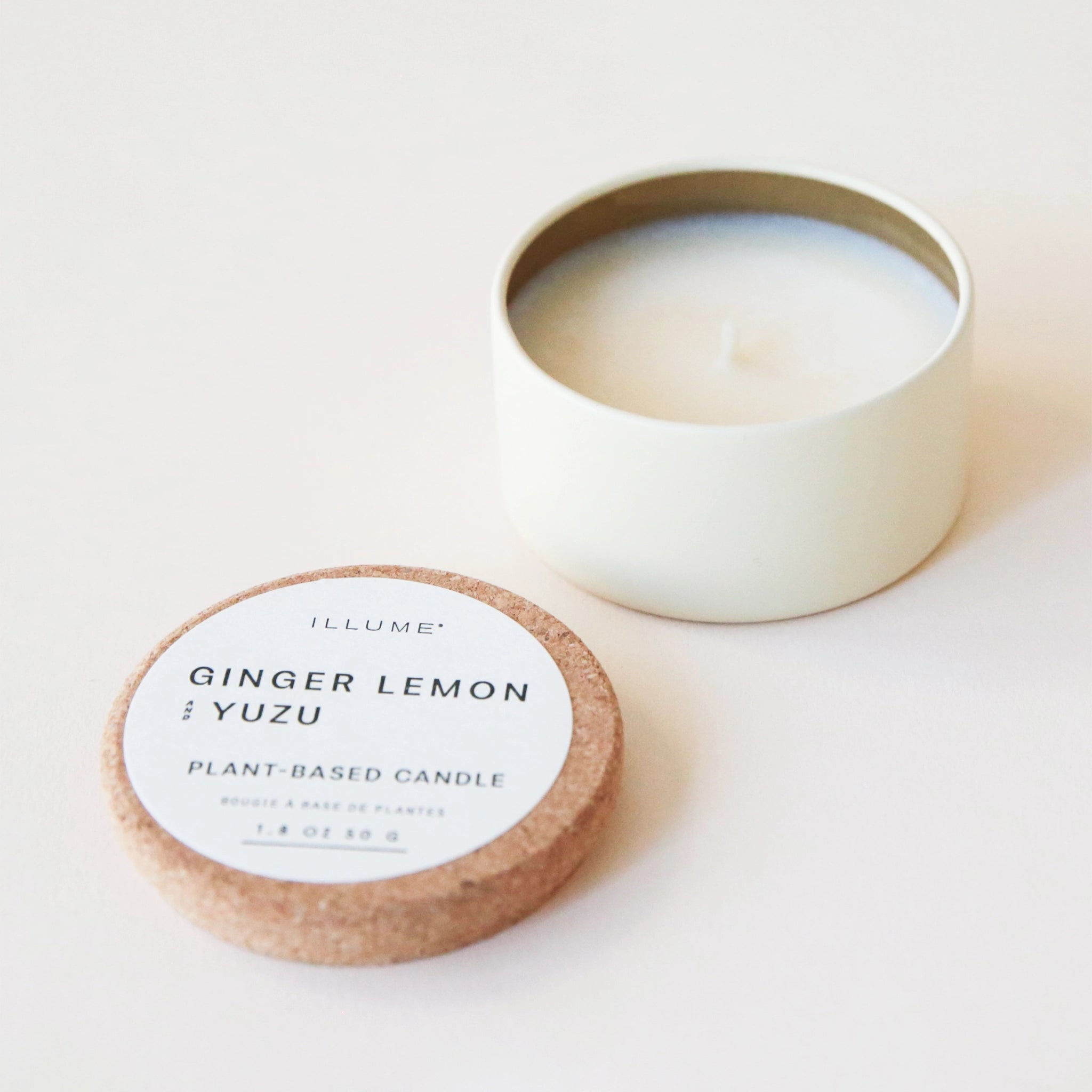 A cream tin candle with white wax and a cork lid that reads, "Ginger Lemon & Yuzu Plant Based Candle" in black letters.