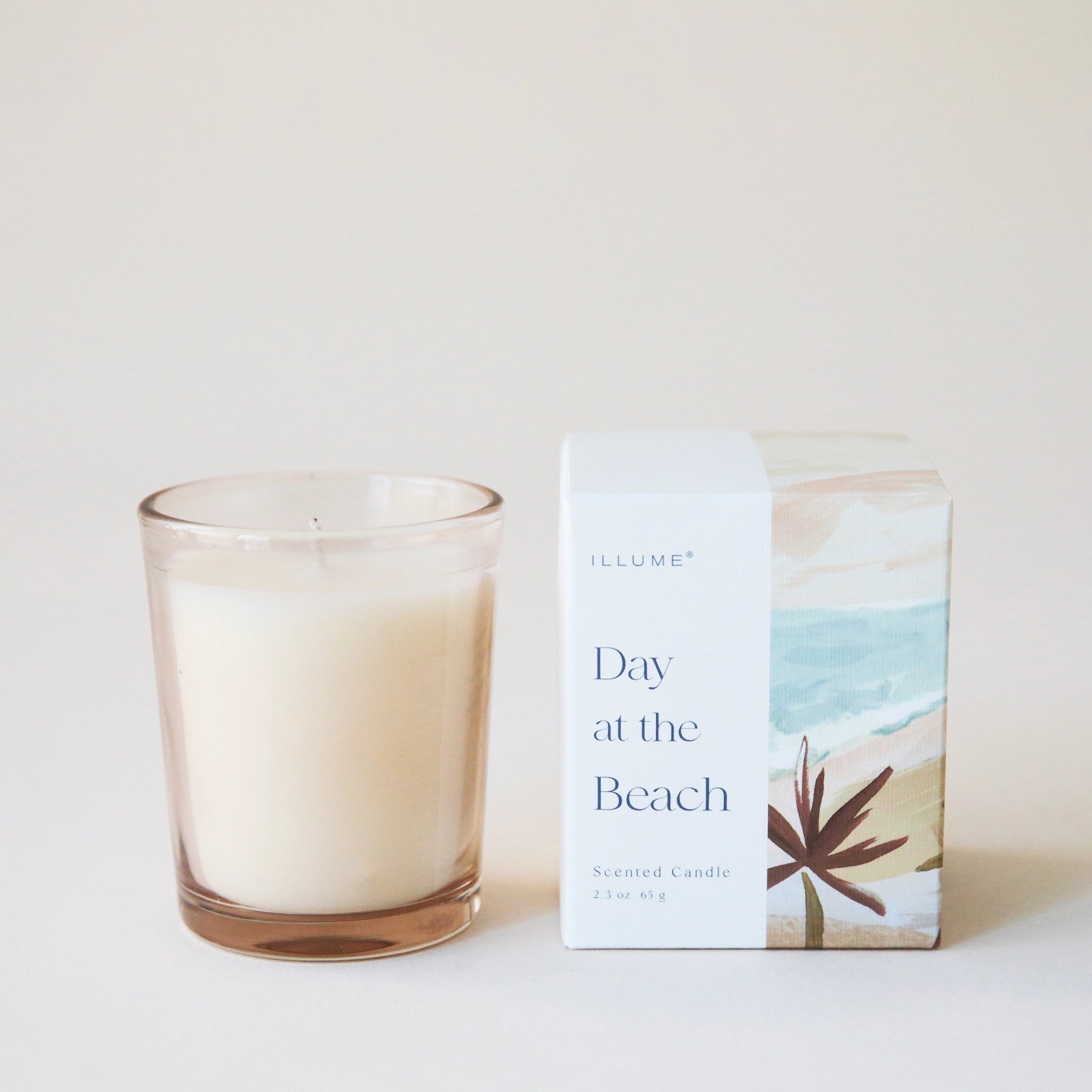 A tan glass jar candle with white wax inside alongside the packaging it comes in that features a white box as well as an illustrated beach scene.