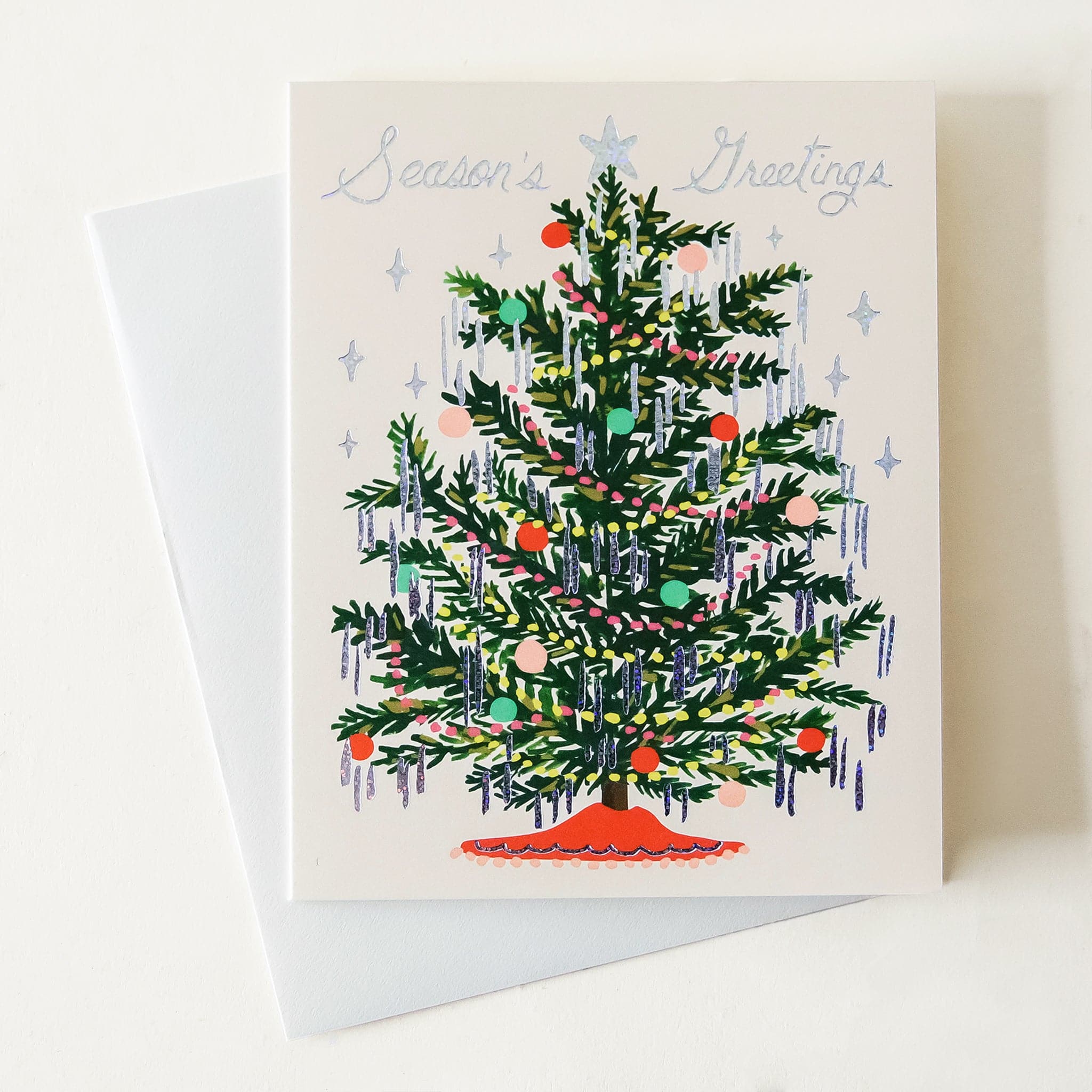 A cream colored card sits against a white background. On the front of the card there is a drawing of a dark green Christmas tree. There are yellow and red Christmas lights up and down the tree. There are green, red and pink ornaments spread out all over the tree. Silver tinsel is hanging on the end of each tree branch. At the bottom is a red tree skirt. At the top is a sliver star. Above the tree in silver script it reads ‘season&#39;s greetings.’ The card is accompanied by a white envelope. 