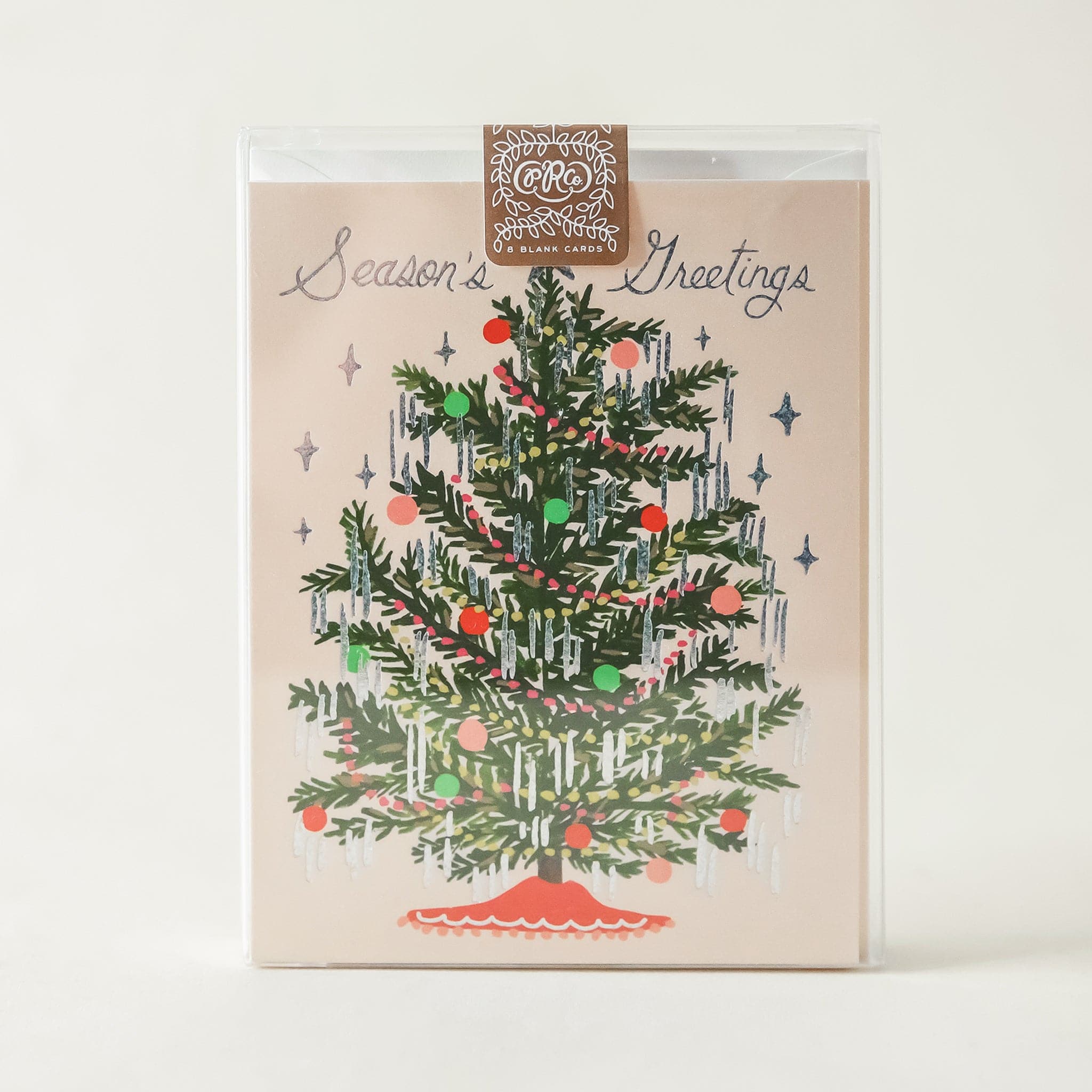 A cream colored card sits inside a clear, plastic case. The case is sitting against a white background. On the front of the card there is a drawing of a dark green Christmas tree. There are yellow and red Christmas lights up and down the tree. There are green, red and pink ornaments spread out all over the tree. Silver tinsel is hanging on the end of each tree branch. At the bottom is a red tree skirt. At the top is a sliver star. Above the tree in silver script it reads ‘season&#39;s greetings.’
