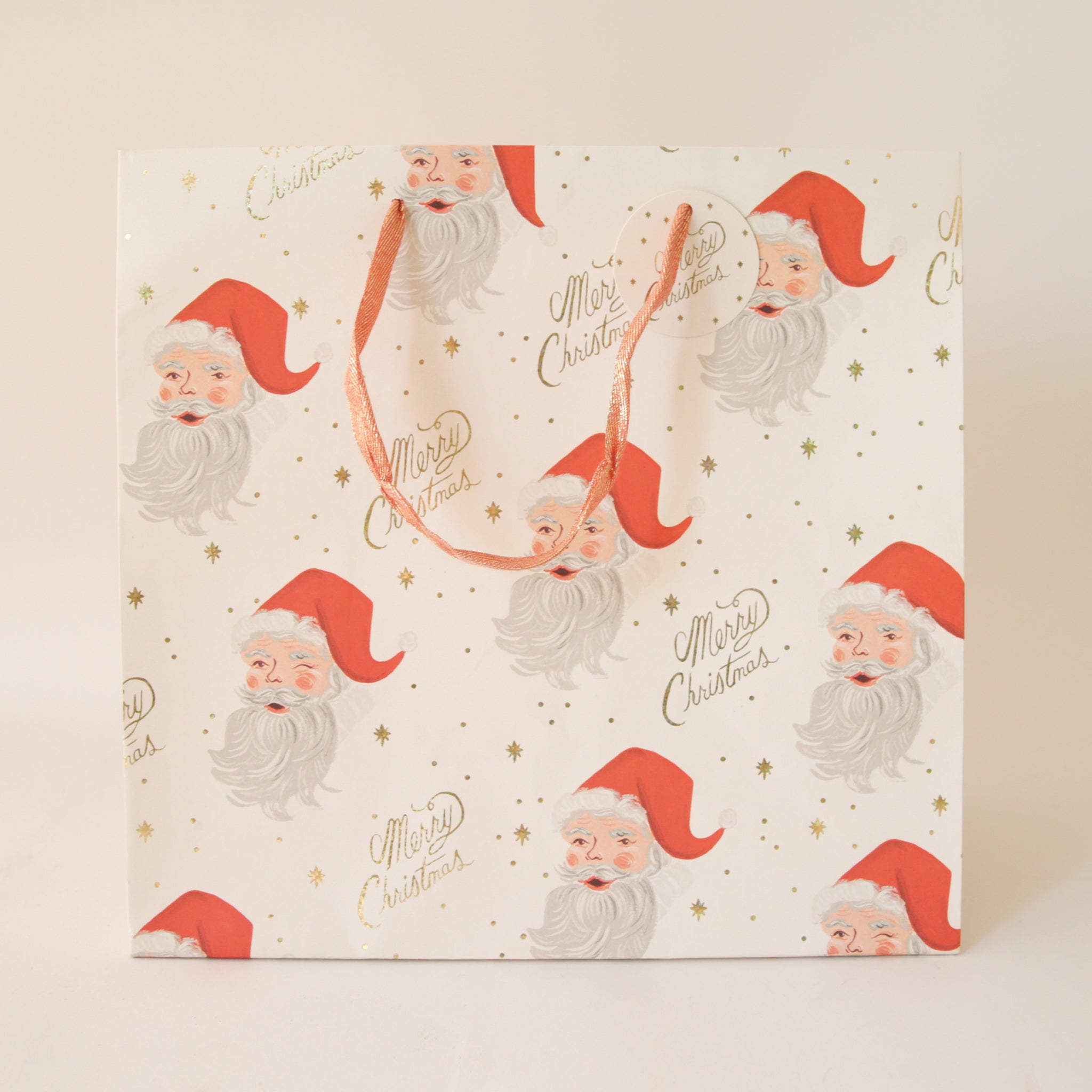 On a tan background an ivory gift bag with a red Santa design along with gold text that reads, &quot;Merry Christmas&quot;.