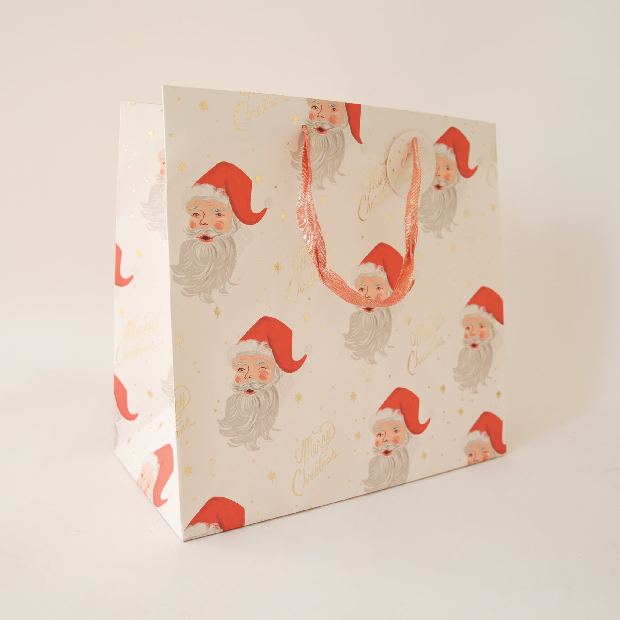 On a tan background an ivory gift bag with a red Santa design along with gold text that reads, &quot;Merry Christmas&quot;.