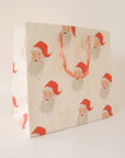 On a tan background an ivory gift bag with a red Santa design along with gold text that reads, "Merry Christmas".