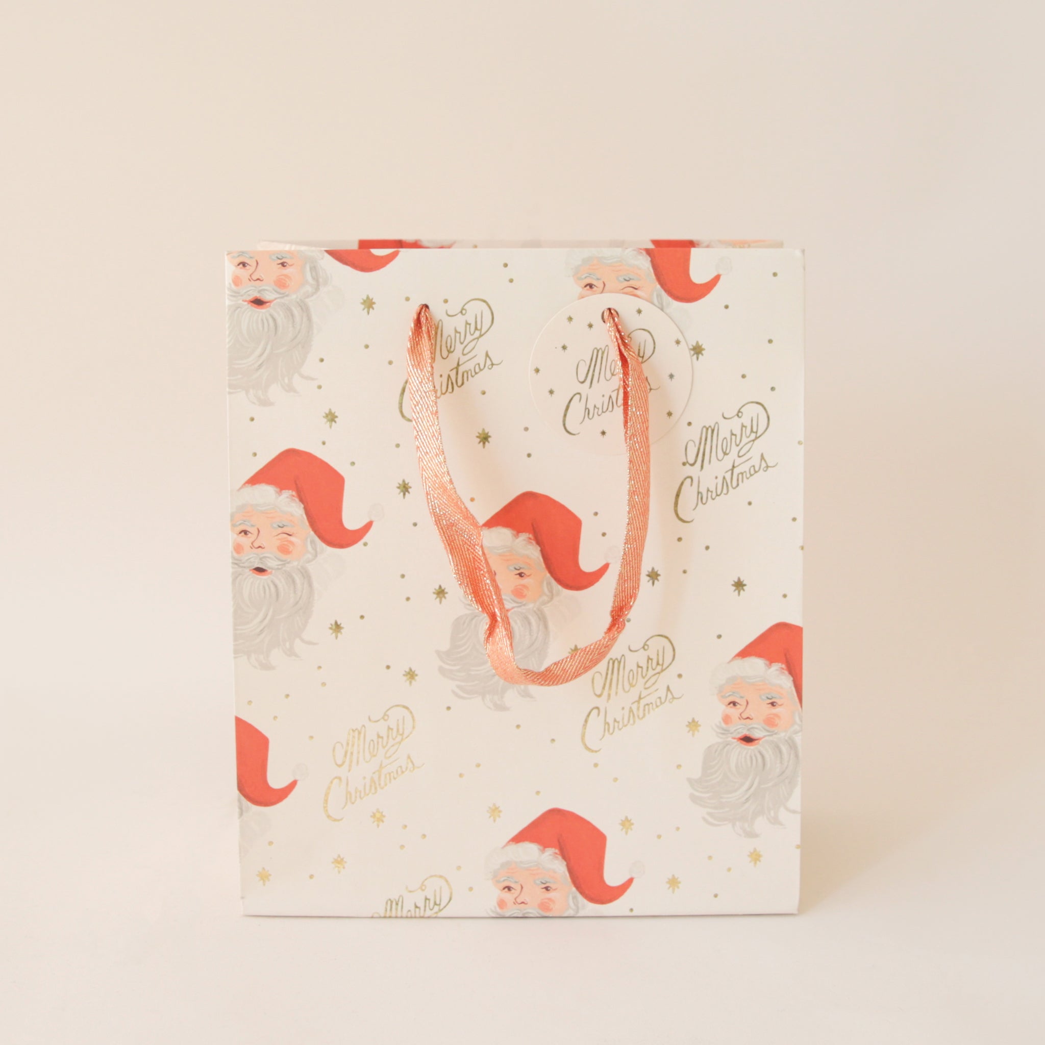 On a tan background an ivory gift bag with a red Santa design along with gold text that reads, &quot;Merry Christmas&quot;.