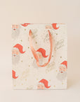 On a tan background an ivory gift bag with a red Santa design along with gold text that reads, "Merry Christmas".