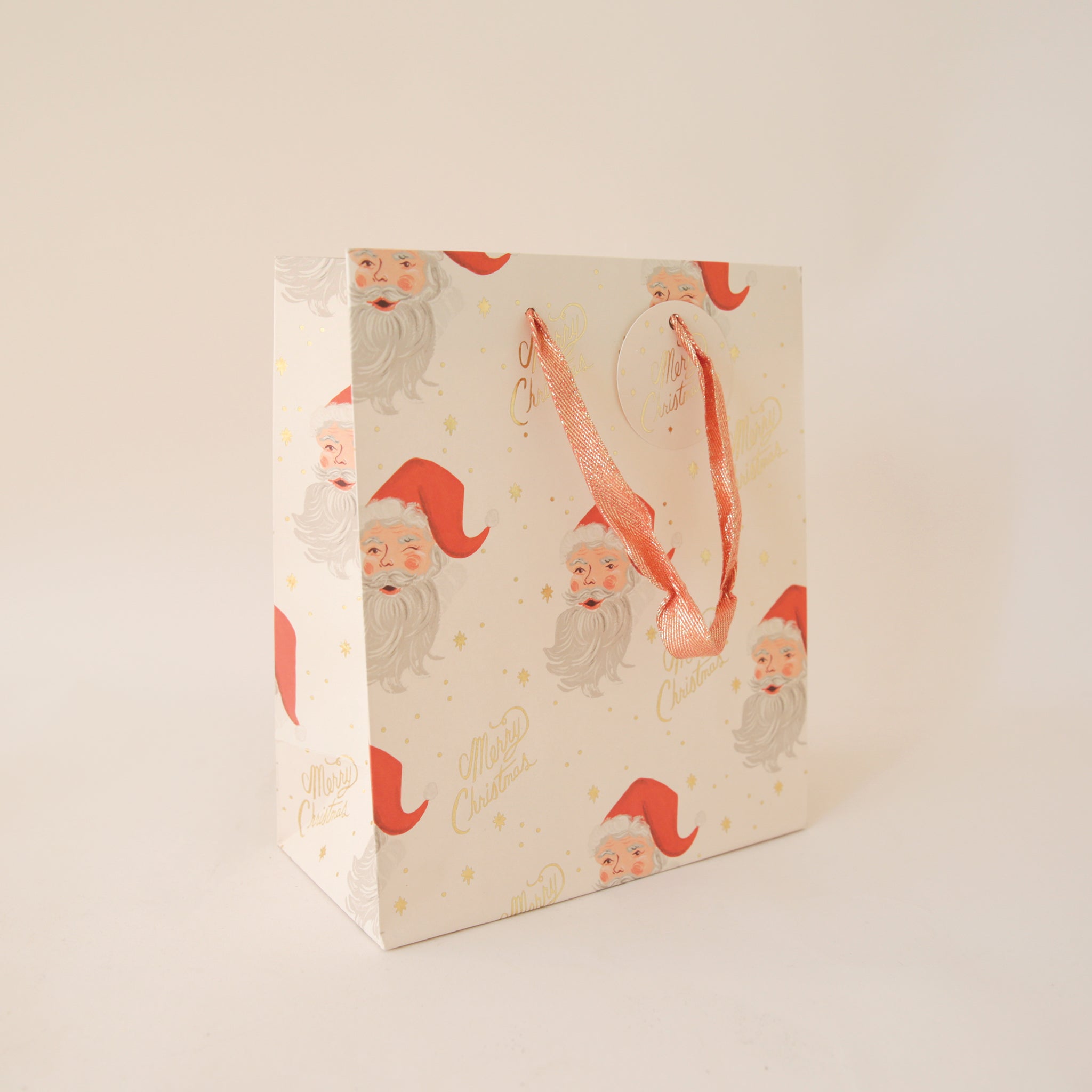 On a tan background an ivory gift bag with a red Santa design along with gold text that reads, &quot;Merry Christmas&quot;.