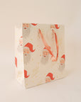 On a tan background an ivory gift bag with a red Santa design along with gold text that reads, "Merry Christmas".