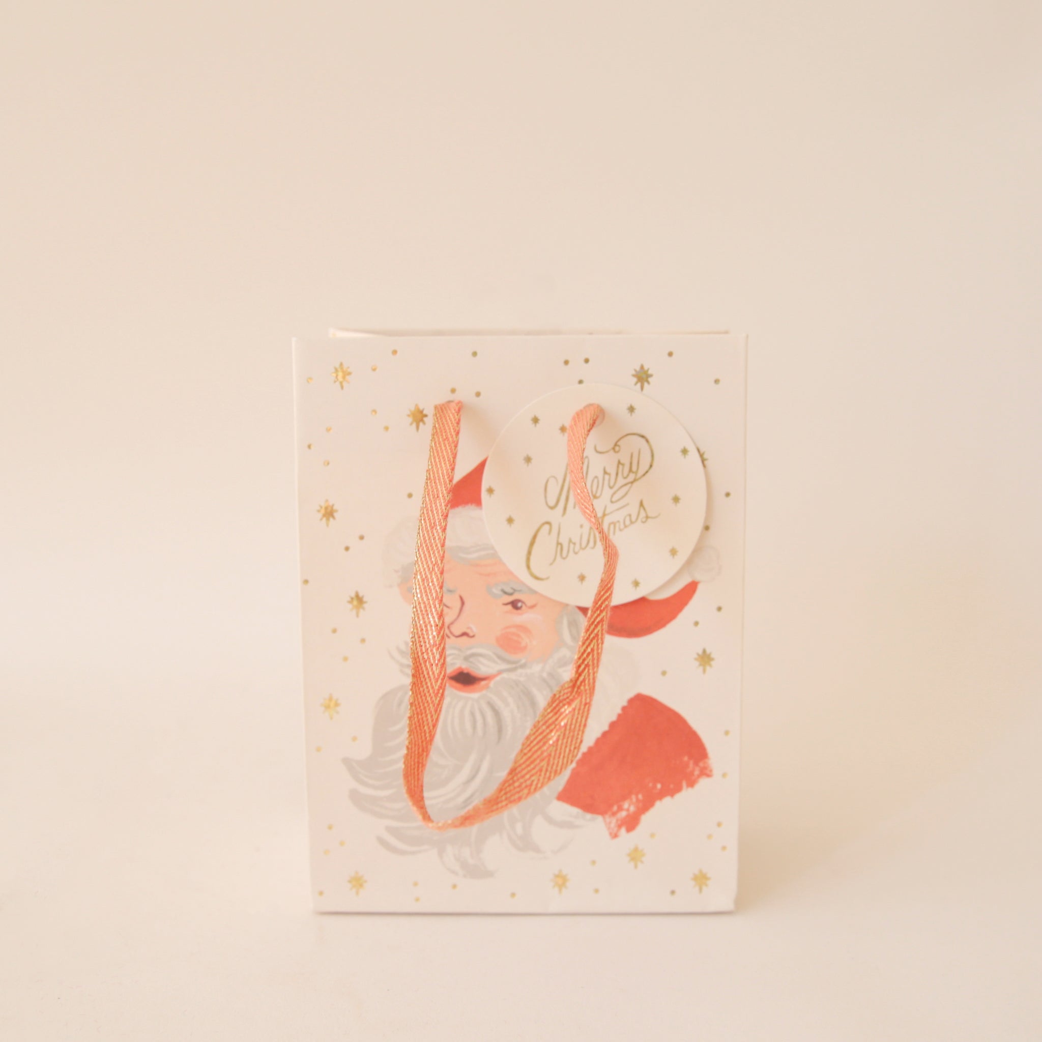 On a tan background an ivory gift bag with a red Santa design along with gold text that reads, &quot;Merry Christmas&quot;.