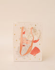 On a tan background an ivory gift bag with a red Santa design along with gold text that reads, "Merry Christmas".