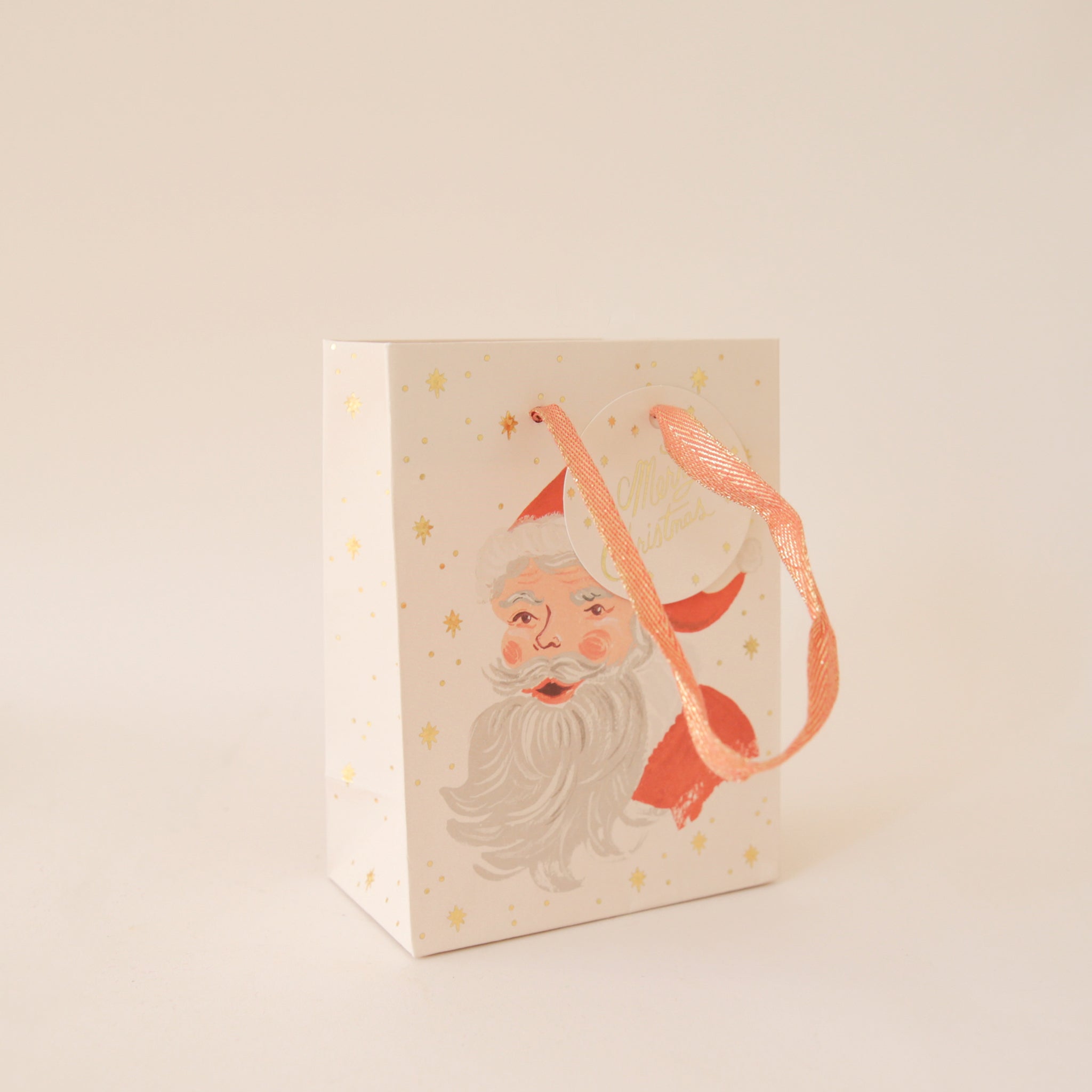On a tan background an ivory gift bag with a red Santa design along with gold text that reads, &quot;Merry Christmas&quot;.