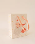 On a tan background an ivory gift bag with a red Santa design along with gold text that reads, "Merry Christmas".