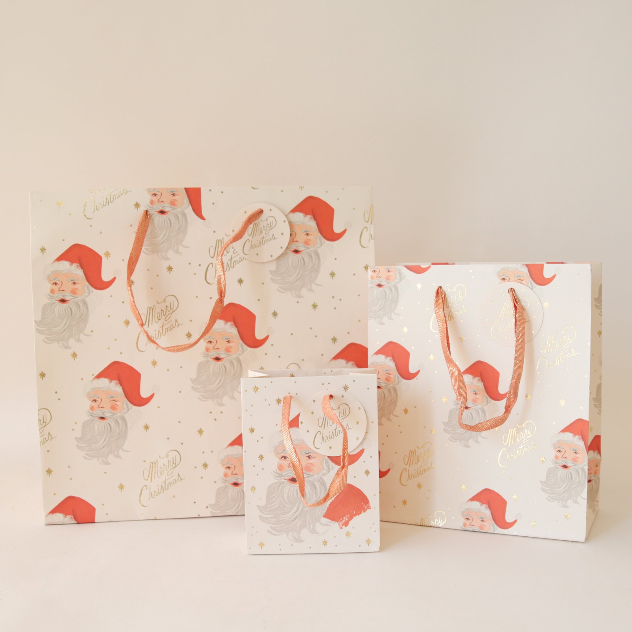 On a tan background is three different sized ivory gift bags with a red Santa design along with gold text that reads, &quot;Merry Christmas&quot;.
