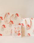 On a tan background is three different sized ivory gift bags with a red Santa design along with gold text that reads, "Merry Christmas".