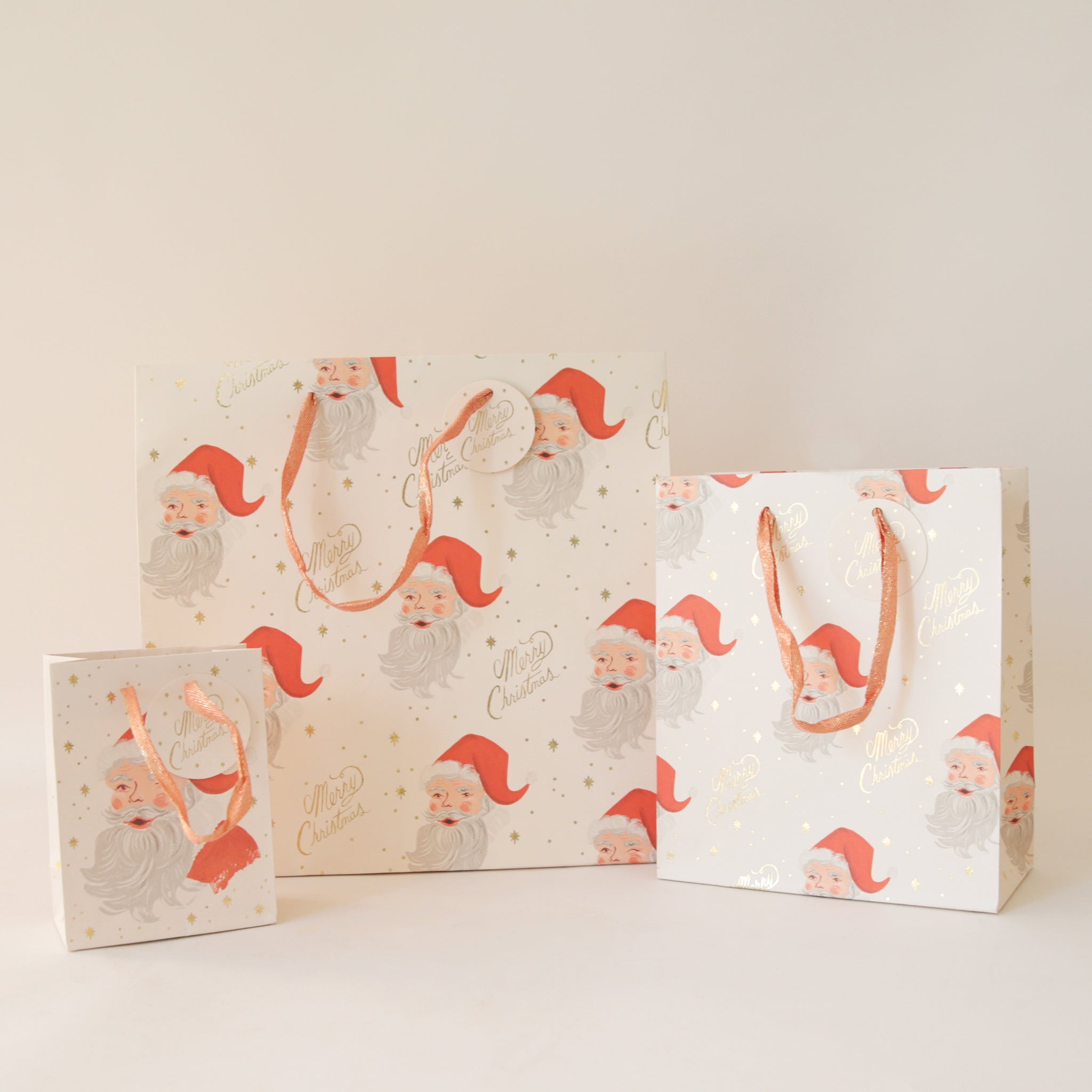 On a tan background is three different sized ivory gift bags with a red Santa design along with gold text that reads, &quot;Merry Christmas&quot;.