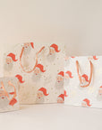 On a tan background is three different sized ivory gift bags with a red Santa design along with gold text that reads, "Merry Christmas".
