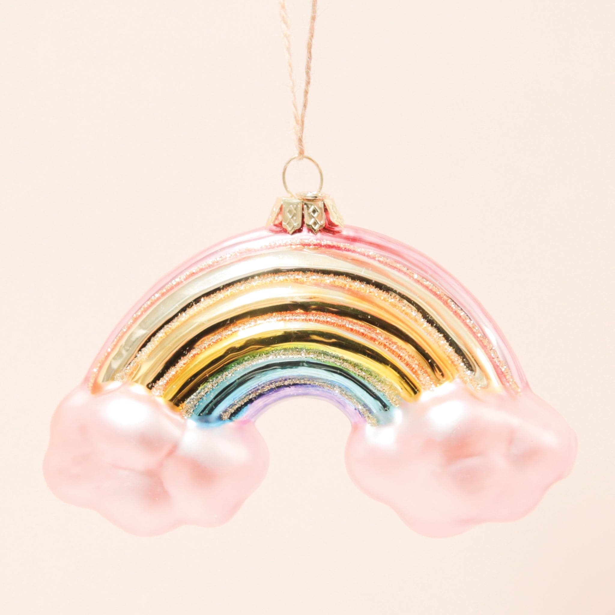 In front of a peachy background is a glass rainbow ornament. There is a light pink cloud on each end of the rainbow. The colors of the rainbow are red, orange, yellow, green, blue and purple. There is gold glitter in between each color. At the top of the ornament is a gold hook with brown twine string.