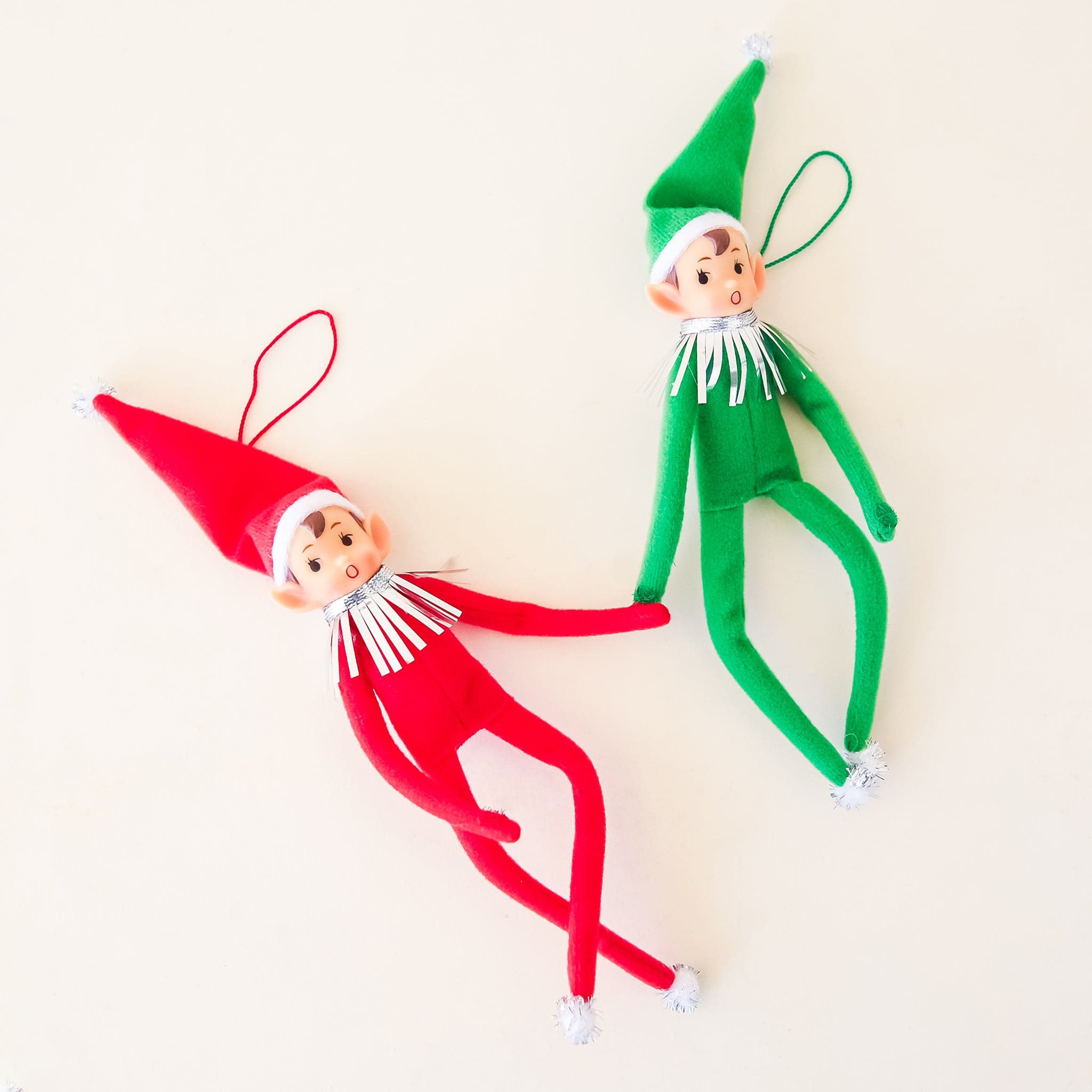 On a cream background is a red elf ornament with long limbs and a tall hat as well as the green version right next to it. 