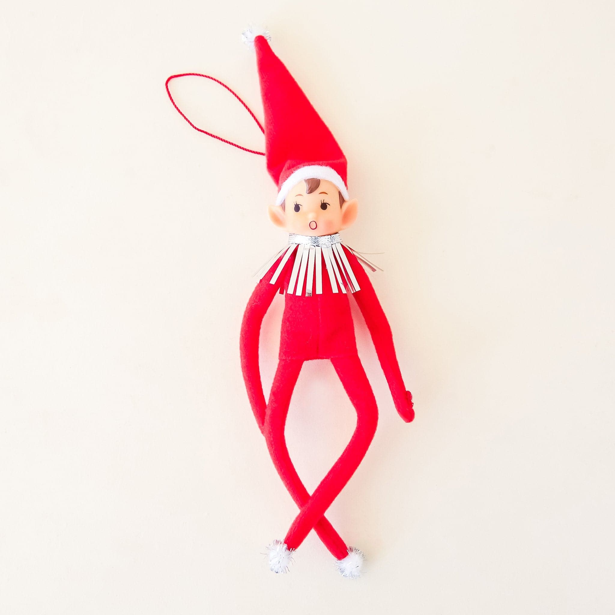 On a cream background is a red elf ornament with bendy arms and legs and a red loop for hanging.