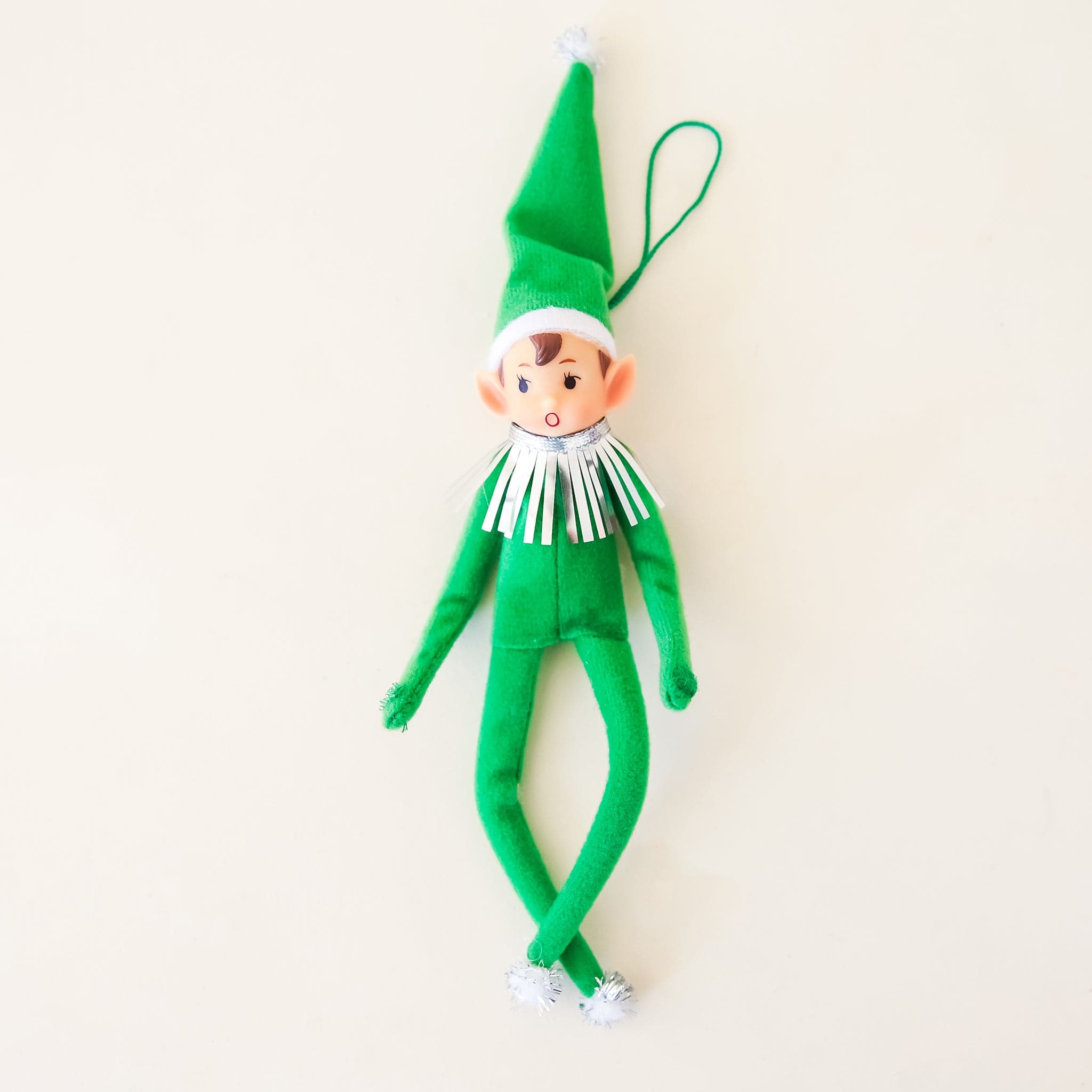 On a cream background is a green elf ornament with bendy arms and legs and a green loop for hanging. 