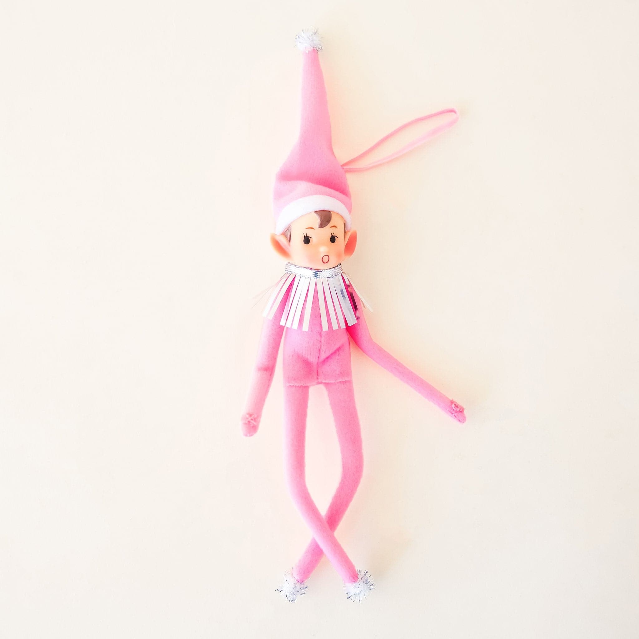 On a cream background is a elf ornament with long arms and legs with a pink suit and hat on as well as a pink string loop for hanging. 