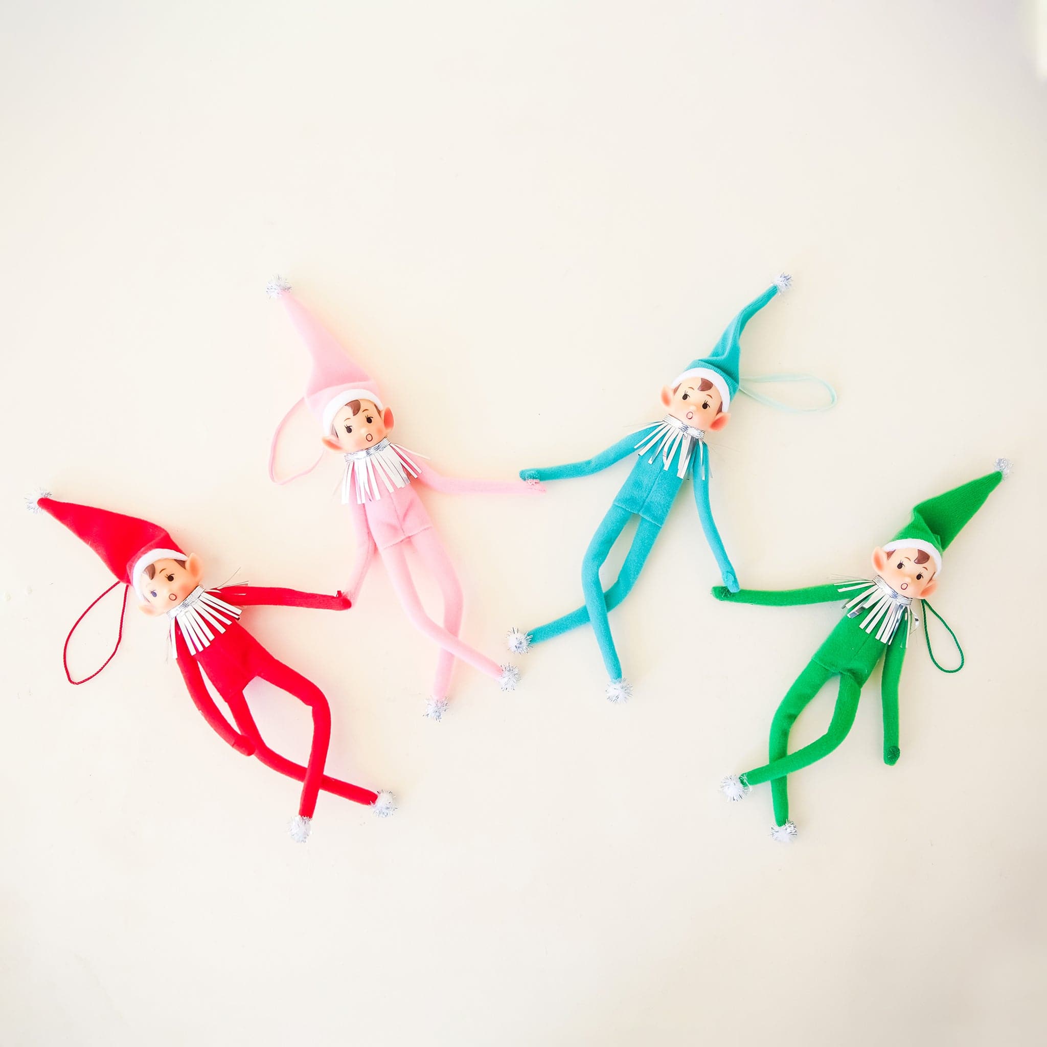 On a cream background is a green elf ornament with bendy arms and legs and a green loop for hanging and photographed next to the other three available colors, red, teal and pink.