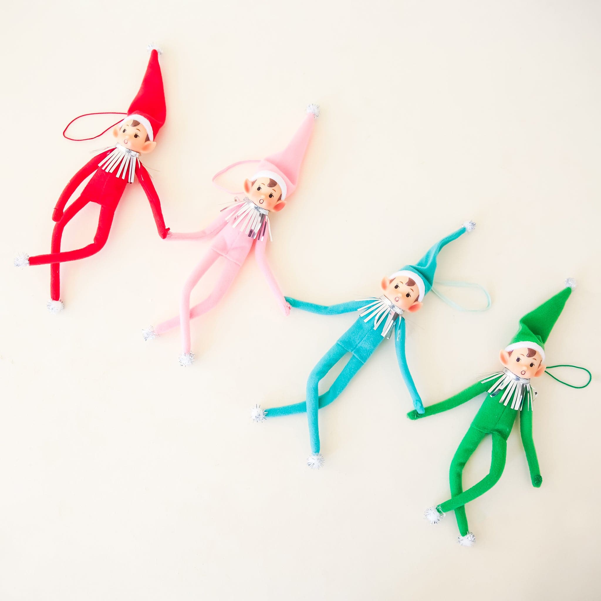 On a cream background is a green elf ornament with bendy arms and legs and a green loop for hanging and photographed next to the other three available colors, red, teal and pink. 
