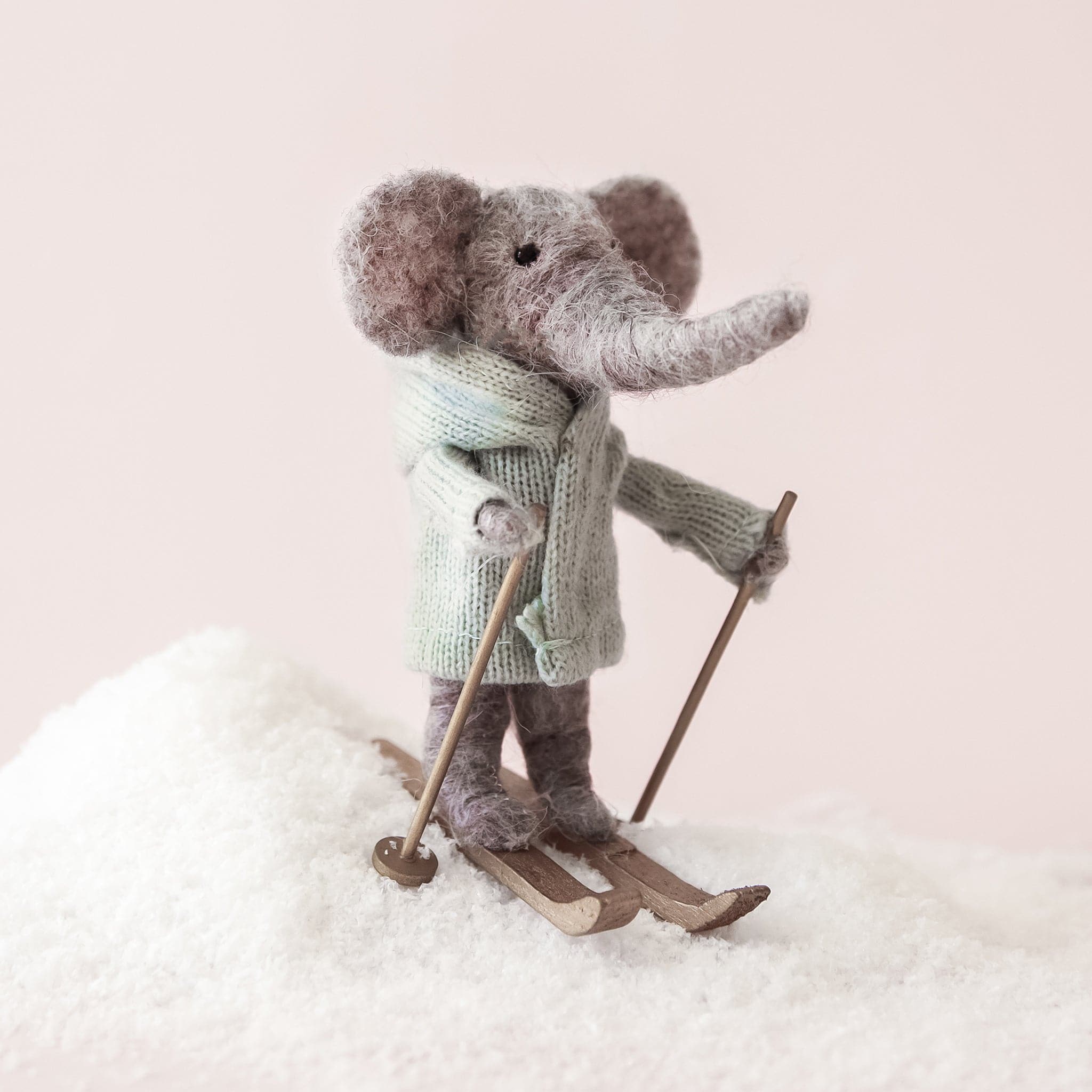 Sitting on a hill of fake snow in front of a pink background is a felt elephant ornament. The elephant is standing upright on his back legs. His feet are on a pair of brown skis and is holding a ski pole in each hand. The elephant is gray with a long, gray trunk and two gray ears. He is wearing a sage green sweater. 