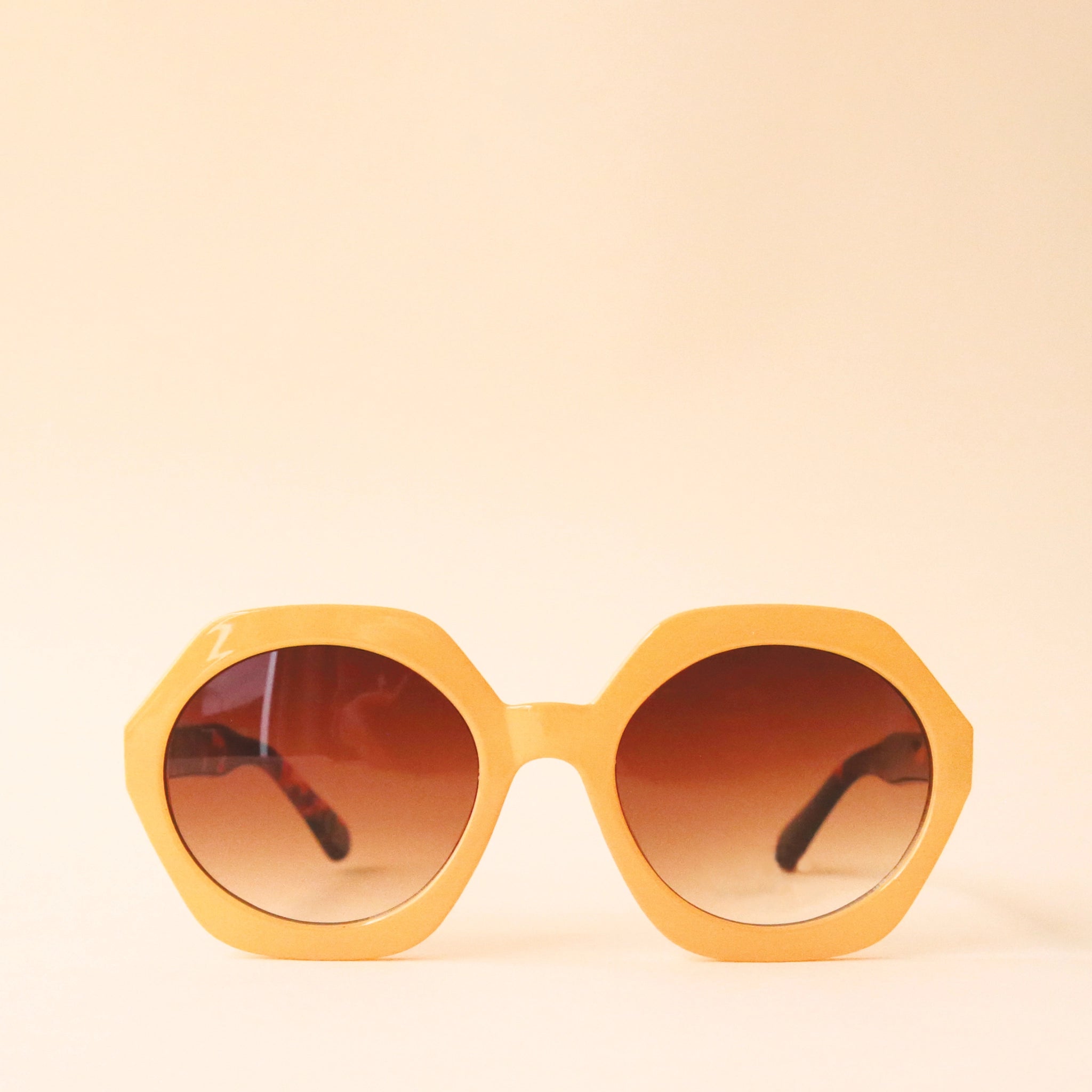 Mustard yellow circular frame sunglasses with a brown lens and tortoise arms photographed in front of a peachy background.