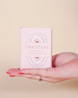 A small pink book that says, "Gratitude Meditations & Inspirations" in light pink on the front cover as well as s geometric illustration above and below the text. 