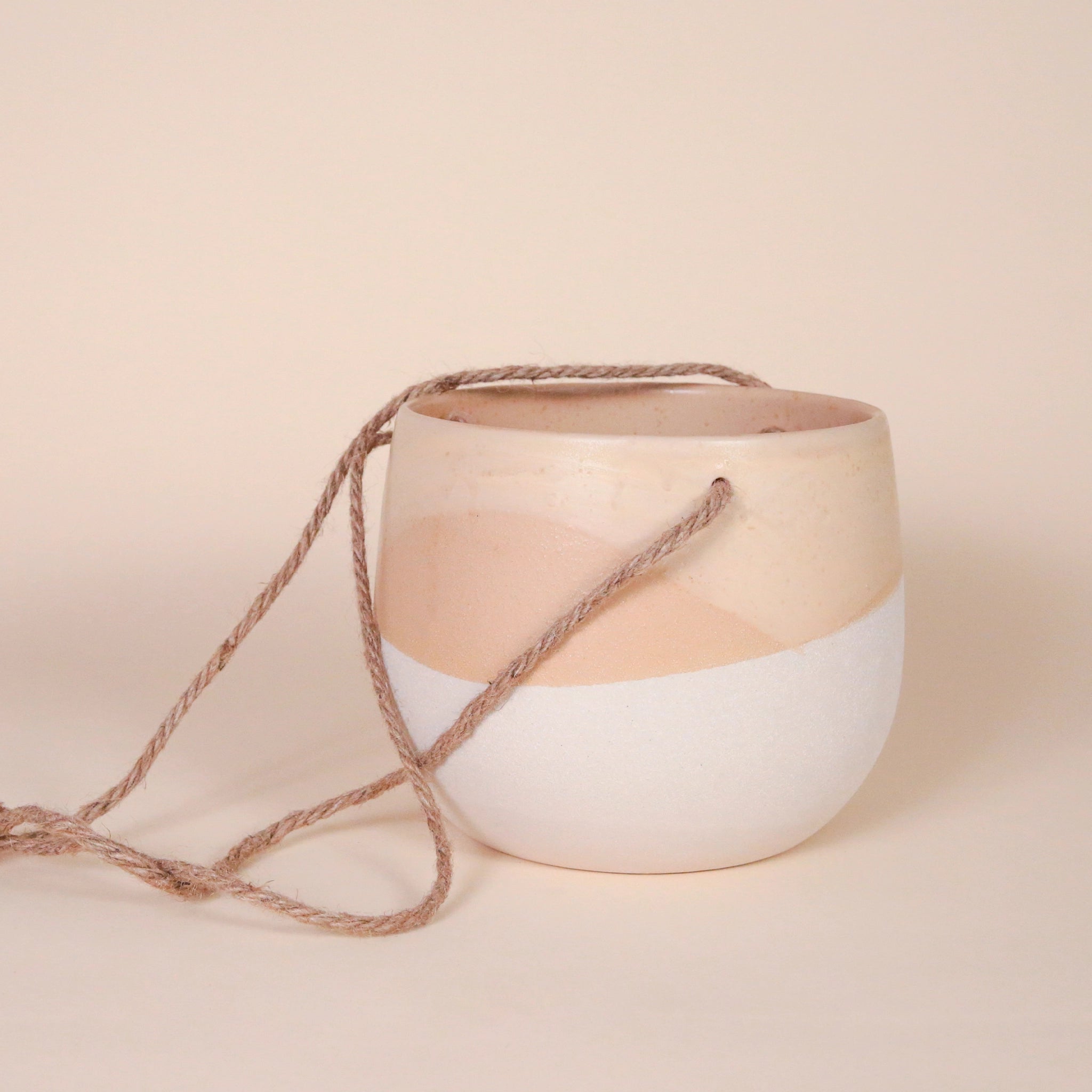 A ceramic circle planter with a wavy design featuring the top half as a neutral tan shade and the bottom half white along with three tan ropes tied together at the top in order to hang.