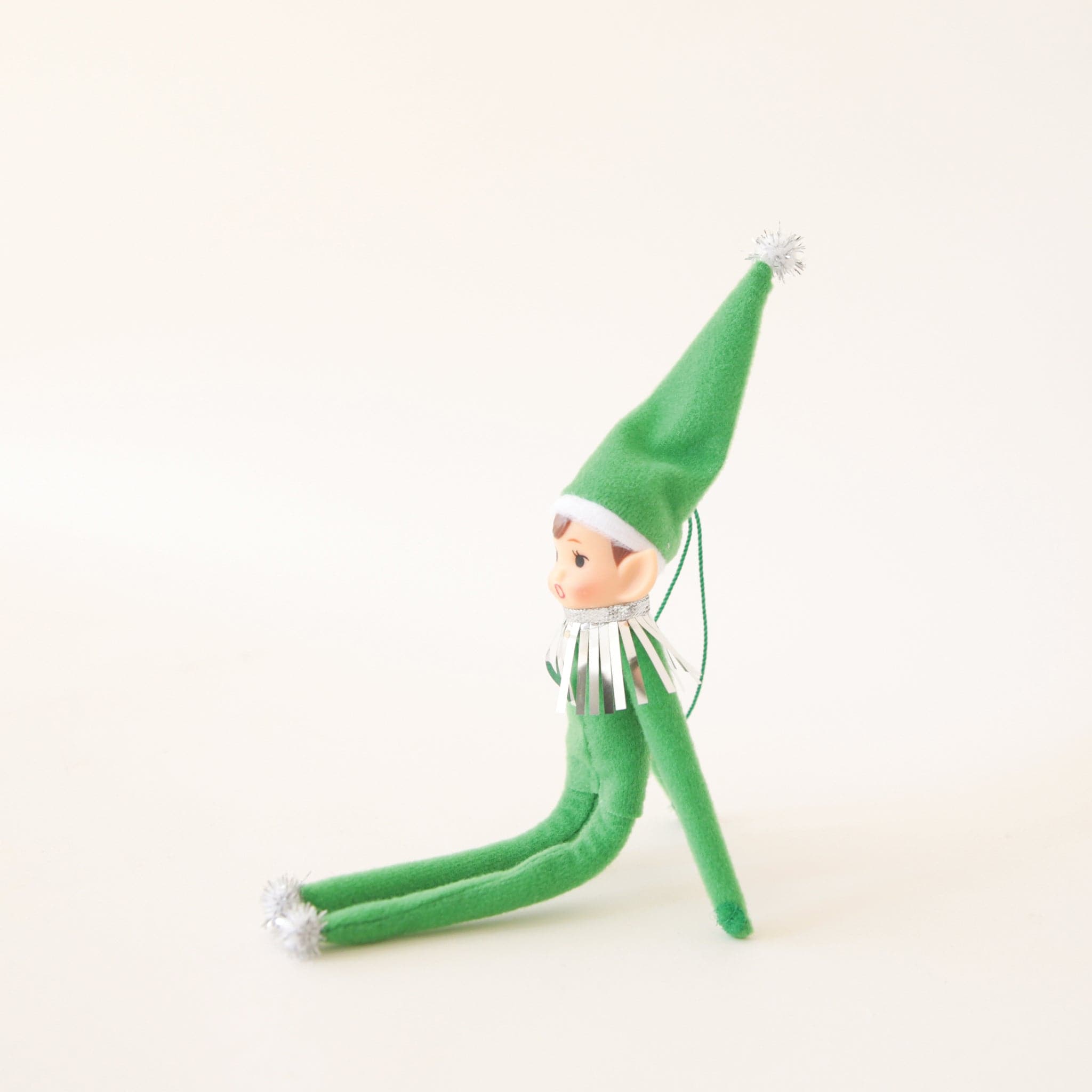 On a cream background is a green elf ornament with bendy arms and legs and a green loop for hanging.