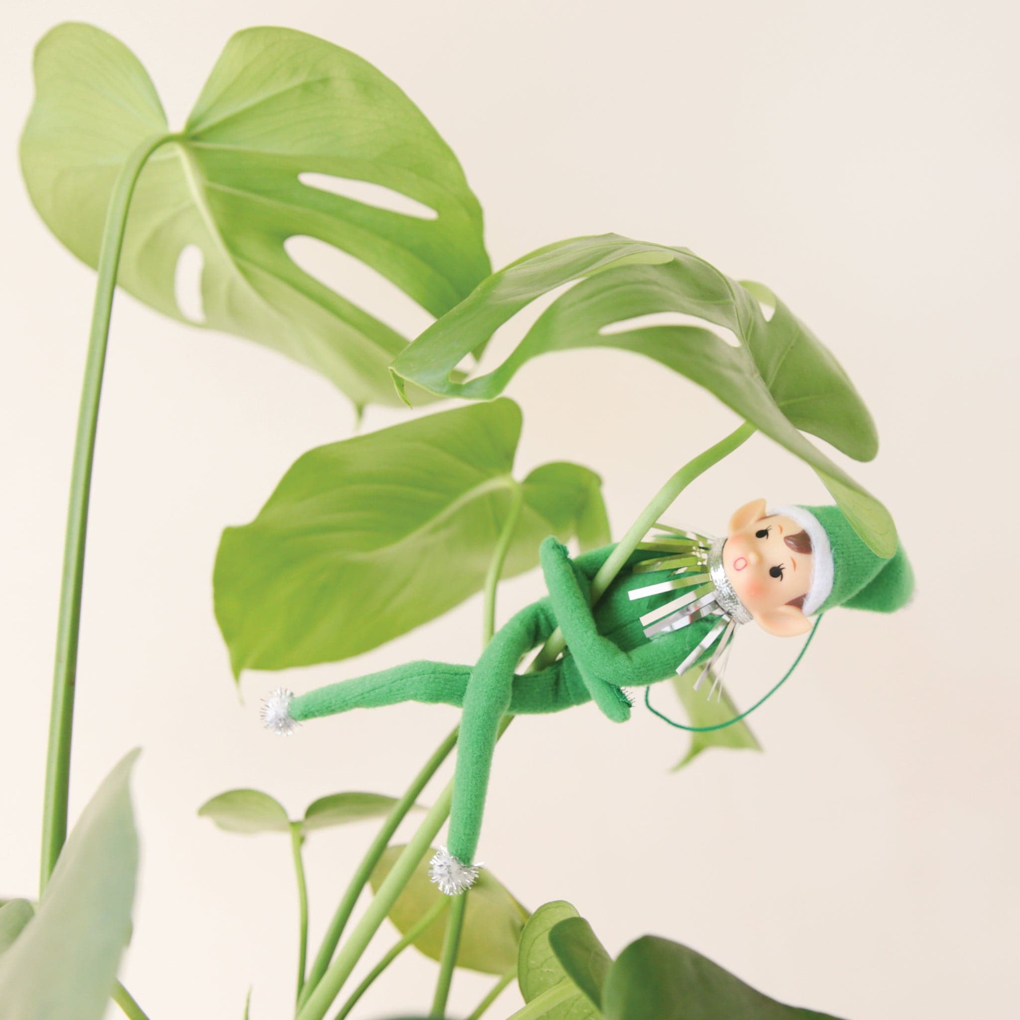 On a cream background is a green elf ornament with bendy arms and legs and a green loop for hanging. In this photo the elf is wrapped around a green house plant. 