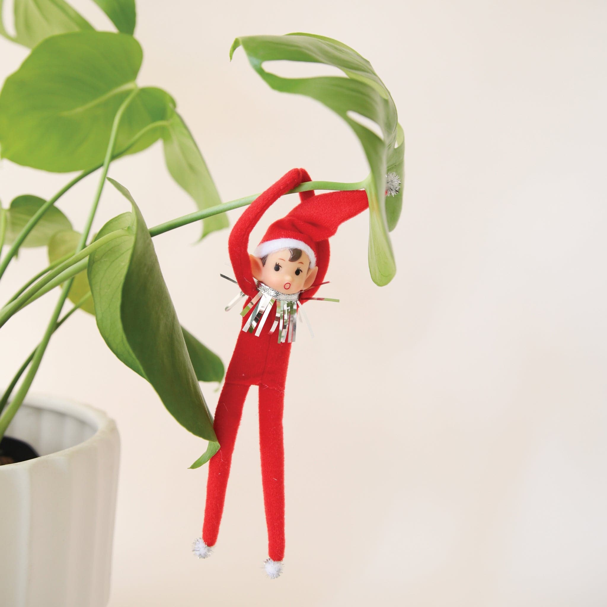 On a cream background is a red elf ornament with bendy arms and legs and a green loop for hanging and is photographed here wrapped around a green house plant.