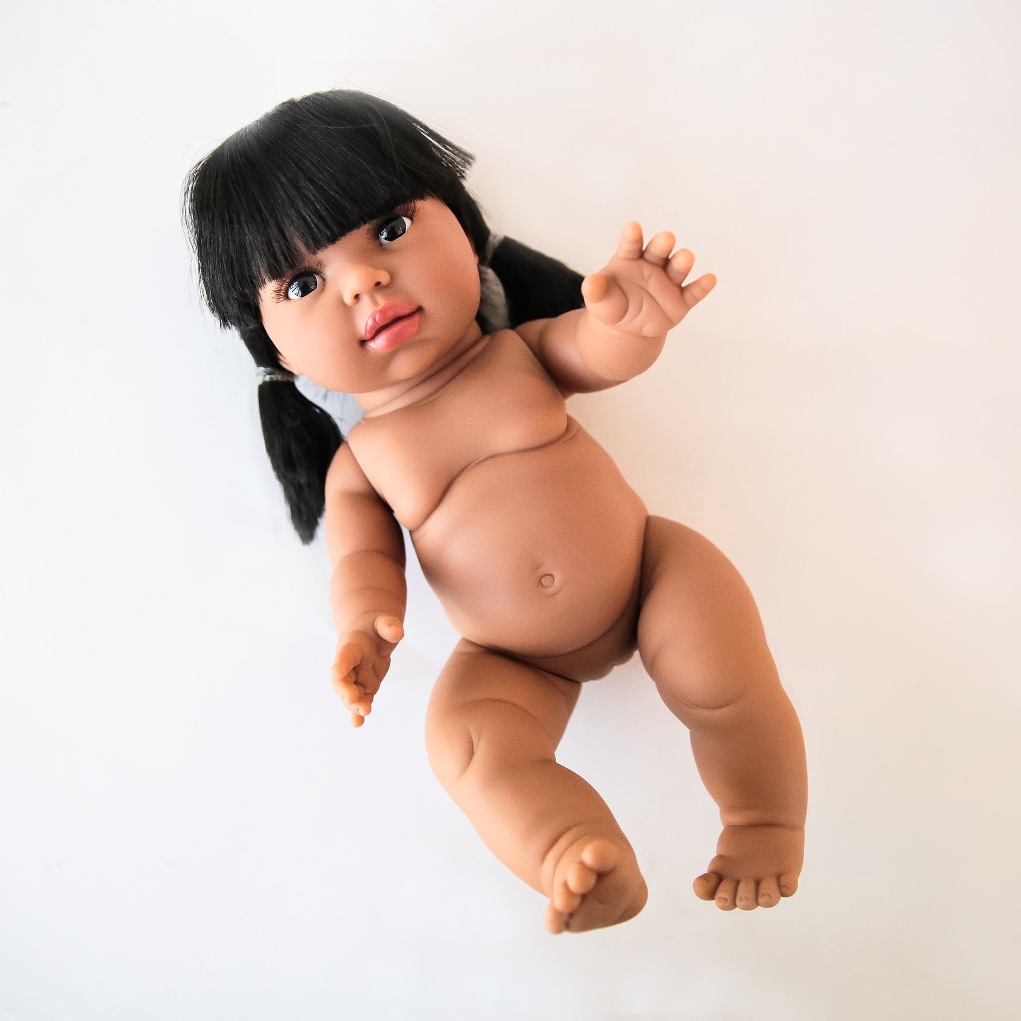 On a white background is a baby girl doll with black pigtails and bangs and brown eyes. 