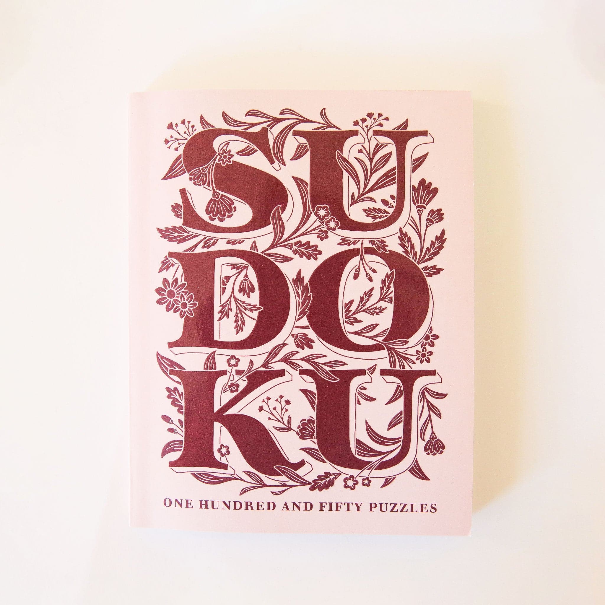 Pastel pink paperback puzzle book titled &#39;Sudoku&#39; in large magenta capital lettering. The letters are accented with delightful floral detailing. Below the large lettering reads &#39;One hundred and fifty puzzles&#39; towards the bottom of the cover. 
