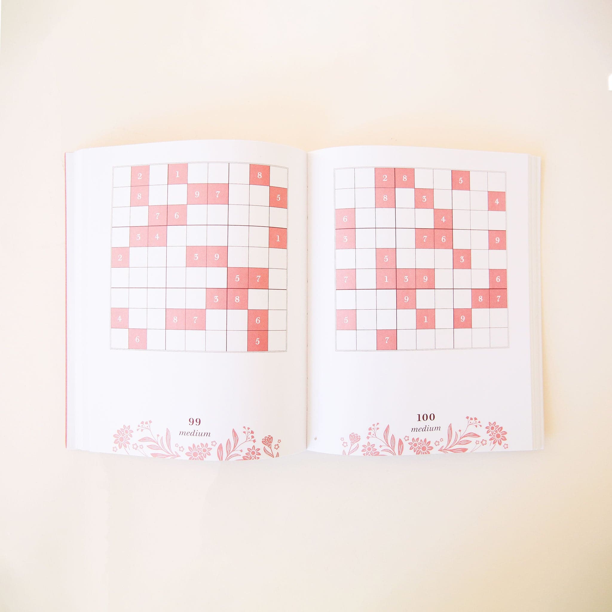 Two open pages filled with sudoku square puzzles. The boxes within the puzzle are blush pink. At the bottom of each page is the number of the puzzle, difficulty level and floral detailing.  